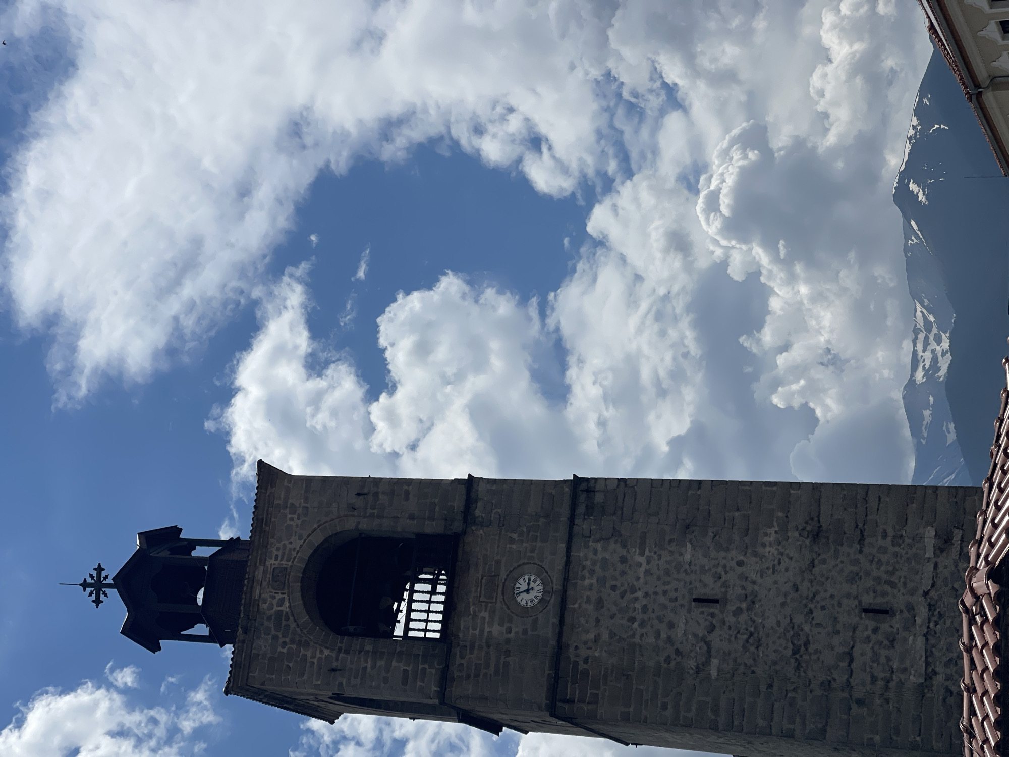 This image captures a stone bell tower under a partly cloudy sky, featuring distant snow-capped mountains and parts of adjacent buildings. The scene is located in the city of Unknown, Unknown.
The central subject is a tall, square-based bell tower constructed from grey, rough-hewn stone blocks, occupying the left half of the frame. Its lower section, largely in shadow, shows two narrow, vertical rectangular slits. Above these, a circular clock face with Roman numerals is embedded in the stone; the hour hand points between XI and XII, and the minute hand points near X, indicating approximately 11:50. Further up, a large, dark arched opening reveals the interior of the belfry, which contains a dark, vertical-slat railing or gate. The very top of the tower features a dark, multi-tiered cupola-like structure, possibly made of wood, crowned with a decorative cross. The upper portions of the tower are significantly darker, suggesting backlighting or deep shadow.
The sky is a vibrant blue, largely filled with voluminous, white cumulus clouds that appear brightly lit from above. Patches of clear blue sky are visible, particularly in the upper right quadrant, where a tiny, dark speck, possibly a bird, can be observed.
In the mid-ground, below the main cloud formation and to the right of the tower, lie several peaks of mountains. These mountains are dark blue-grey and exhibit significant patches of white snow near their summits.
In the bottom left corner, a section of a terracotta-tiled roof with curved tiles is visible. In the bottom right, the top edge of a light-colored building with a dark trim is partially discernible.