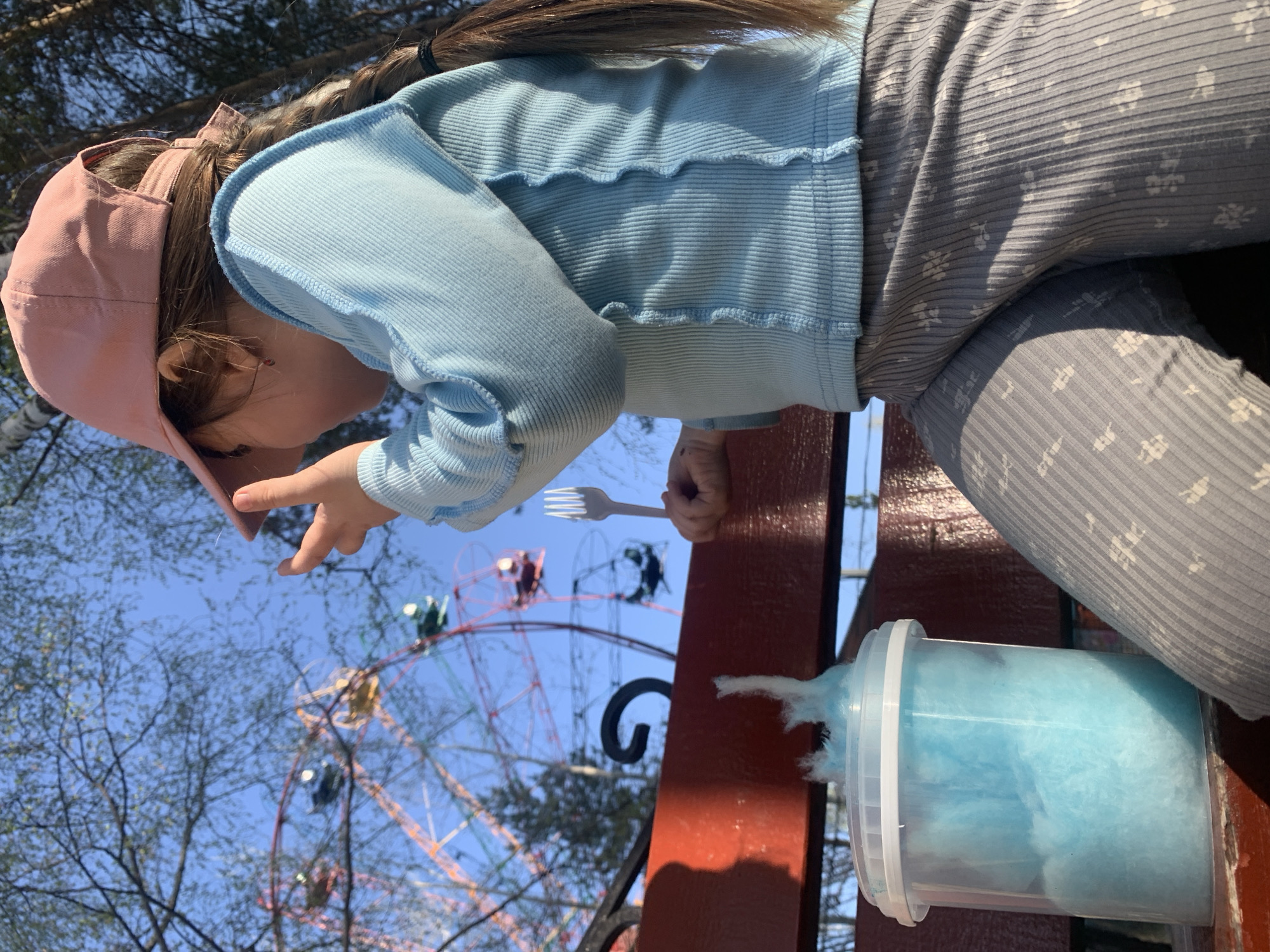 A child wearing a pink cap and a blue shirt is sitting on a red bench at a park near the Ferris wheel. She is holding a plastic fork and reaching out with her other hand. A container of blue cotton candy is next to her on the bench. The location is Chaikovsky, Russia.