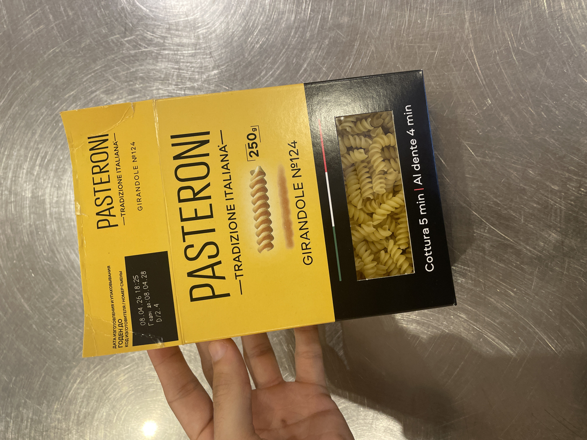 A person's hand is holding a yellow box of pasta. The box has the brand name "PASTERONI" printed in large black letters. Below the brand name, it says "TRADIZIONE ITALIANA" and "GIRANDOLE N°124". There is an image of fusilli pasta on the box, and a small rectangle indicating "250g". A window in the box reveals the actual pasta inside. At the bottom of the box, text reads "Cottura 5 min | Al dente 4 min". A label on the top left of the box has Cyrillic text that translates to "Date of manufacture and packaging", "BEST BY", and "Manufacturer code/shift number". Below this are the dates "08.04.26 18:25" and "BEST BY 08.04.28". The box is placed on a metallic, brushed surface. The lighting suggests an indoor environment, possibly a kitchen or pantry, and the surface indicates a countertop. There is no discernible information in the image to indicate a specific location in the city of Oryol, Russia.