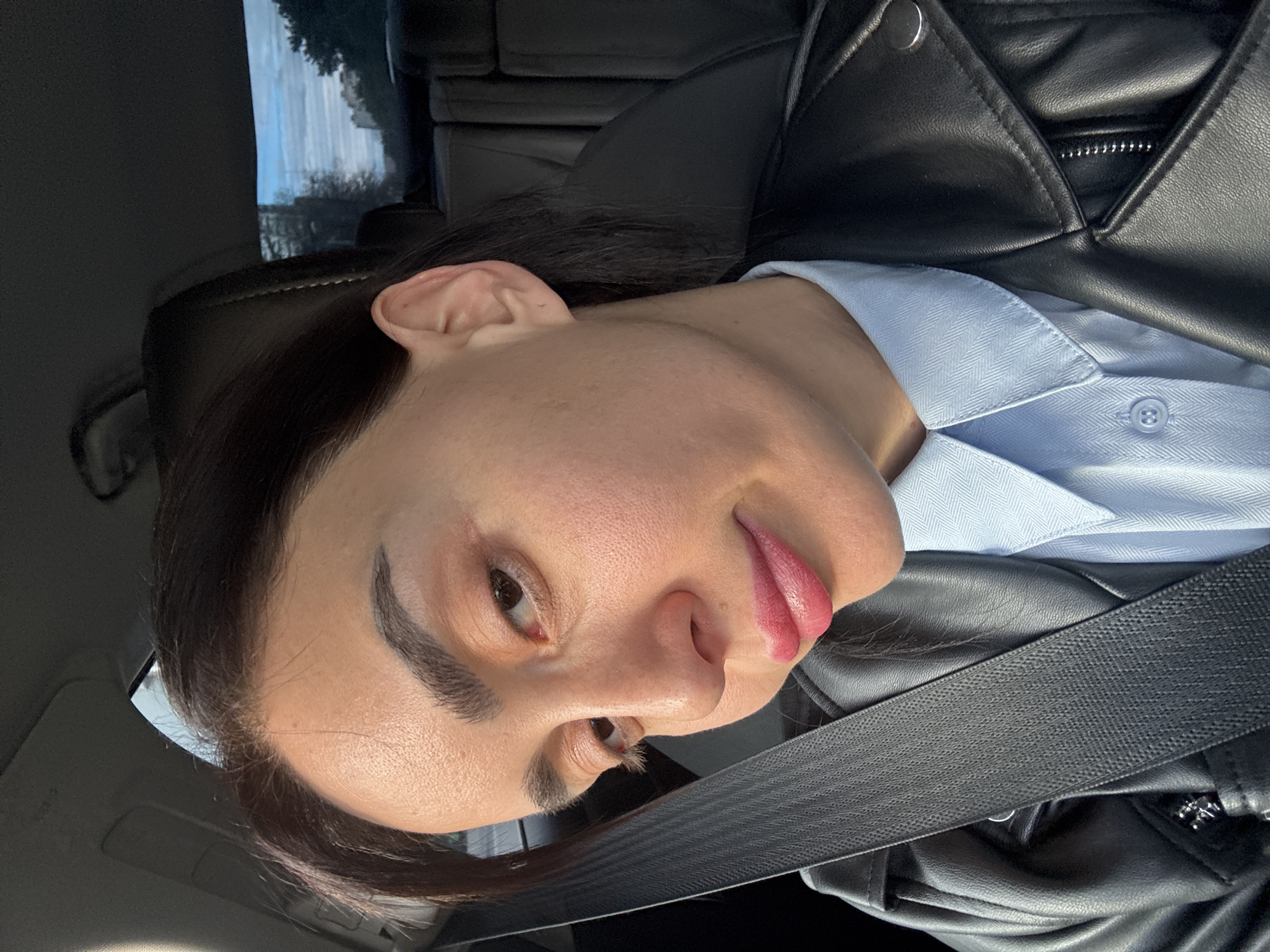 The image depicts a woman in a car, wearing a light blue shirt and a black leather jacket. She is wearing a seatbelt. The background shows an out-of-focus view of trees and buildings, suggesting a location in a city. The details do not allow determining an exact location in Tbilisi.