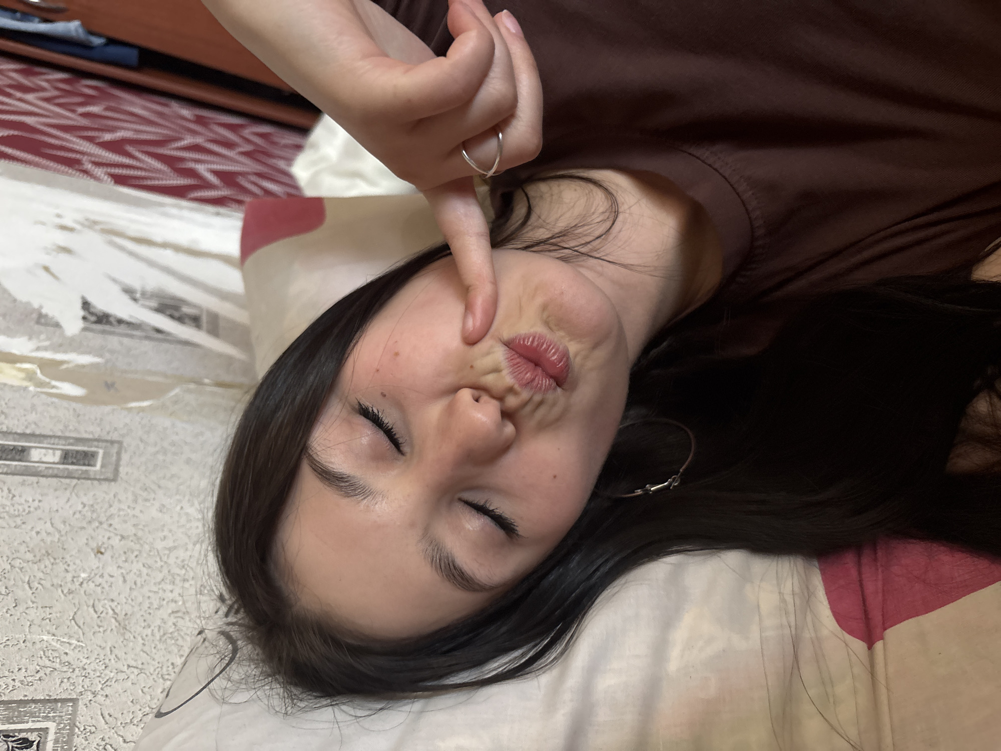 A young woman with dark hair is lying down, eyes closed, making a pouting face while holding her finger to her face. She is wearing a brown shirt and a ring on her finger. She is lying on a patterned pillow and bedspread. The location is not discernible from the image.