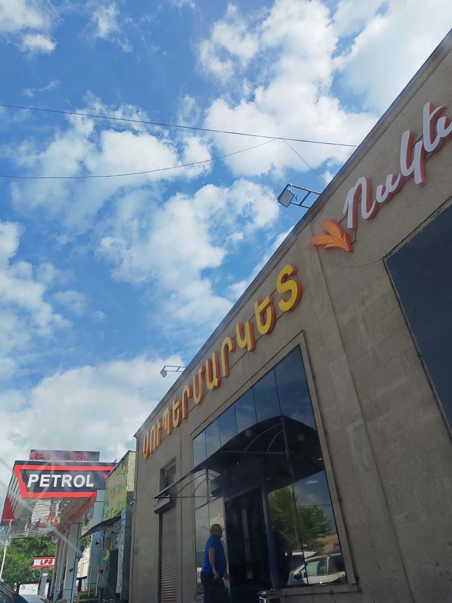 The image shows a building with Armenian lettering and a gas station in Vanadzor, Armenia. The building has a sign with orange and white letters that reads "Նուկե" in Armenian. Below this sign is another sign with yellow letters that reads "ՍՈՒՊԵՐՄԱՐԿԵՏ" in Armenian. To the left of the building is a gas station with a "PETROL" sign and an "LPG" sign. There is a person in a blue shirt walking past the entrance of the building. The sky is blue with white clouds. The image appears to have been taken from a car.