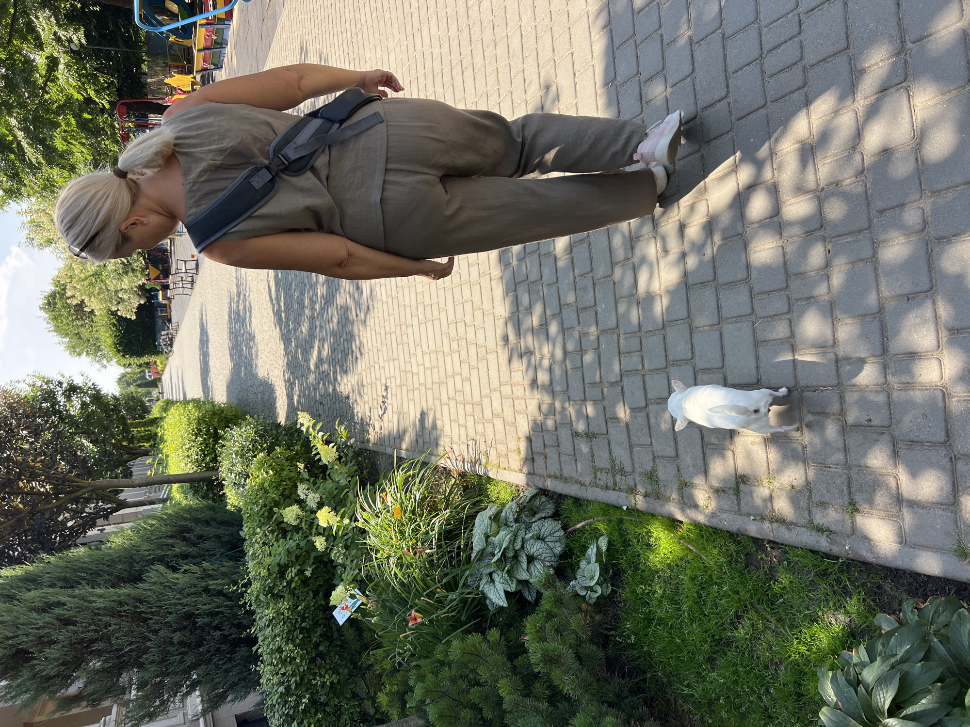 A woman with blonde hair pulled back in a ponytail is walking away from the camera on a paved walkway. She is wearing a sleeveless olive-green jumpsuit and pink sneakers. A dark gray bag is slung across her back. A small white dog is walking on the walkway in front of her, facing away from the camera. The walkway is lined with green bushes, trees, and flowers. In the background, there are more trees and some playground equipment. The scene appears to be in Fontanka, Ukraine.
