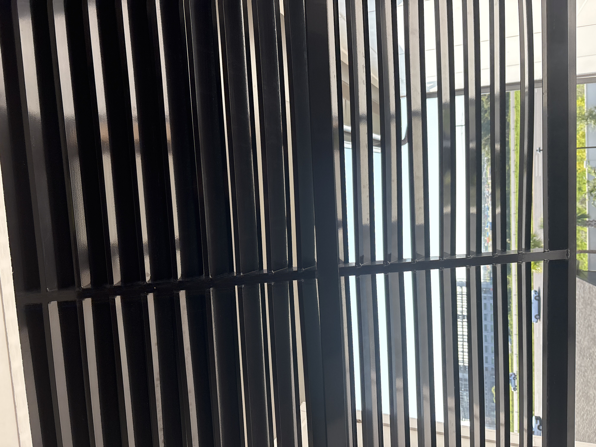 The image shows a view through horizontal, dark-colored blinds. The blinds are tilted, allowing a view of the outdoors. In the distance, a building with multiple floors and reflective windows can be seen, suggesting an urban or commercial area. A road with several cars is visible in the middle ground. The sky appears bright, indicating daylight. The overall impression is of a window in a building looking out onto a street scene with some architectural structures. The provided location information identifies the scene as Palmetto, United States.