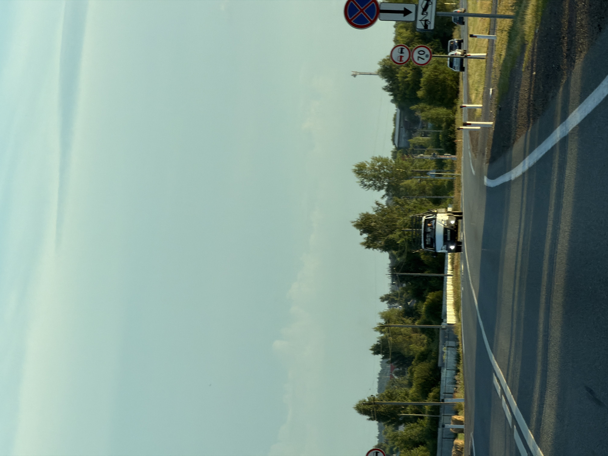 The image shows a two-lane road in a rural setting, likely near Togashevo, Russia, given the provided context. The road curves to the right, with a white lane divider. On the right side of the road, there are several traffic signs. One sign, at the top, is a circular red sign with a blue cross inside, indicating no stopping. Below it are two circular signs, one with two cars side-by-side indicating a passing zone, and another with "70" suggesting a speed limit. Further down, there is a sign depicting a tow truck, likely indicating a towing zone or service. The road is bordered by a grassy shoulder and a low white fence. Beyond the fence, there are trees and some houses visible in the distance. A white van is driving away from the camera on the right lane, and two cars are parked on the right side of the road. The sky is a pale blue with scattered clouds, and the sunlight suggests it is either early morning or late afternoon. The overall scene depicts a typical roadside environment in a suburban or rural area.