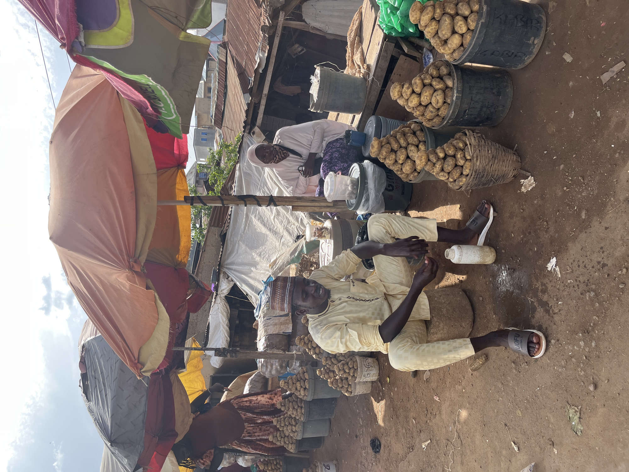This image depicts an outdoor market scene in Jos, Nigeria, likely a roadside stall. Two individuals are visible, both appearing to be vendors. In the foreground, a man wearing a light yellow traditional outfit and a woven hat is seated on a cylindrical object. He is looking downwards and to his right, with his hands resting in his lap. In the background, a woman in a light pink head covering and a patterned garment is seated behind a table laden with goods, including buckets and plastic containers. Several large piles of potatoes are displayed in buckets and a woven basket, suggesting they are for sale. Various market stalls with umbrellas and corrugated metal roofs are visible, indicative of a bustling marketplace environment under a cloudy sky. The ground is dirt, with scattered debris, typical of an informal market setting.