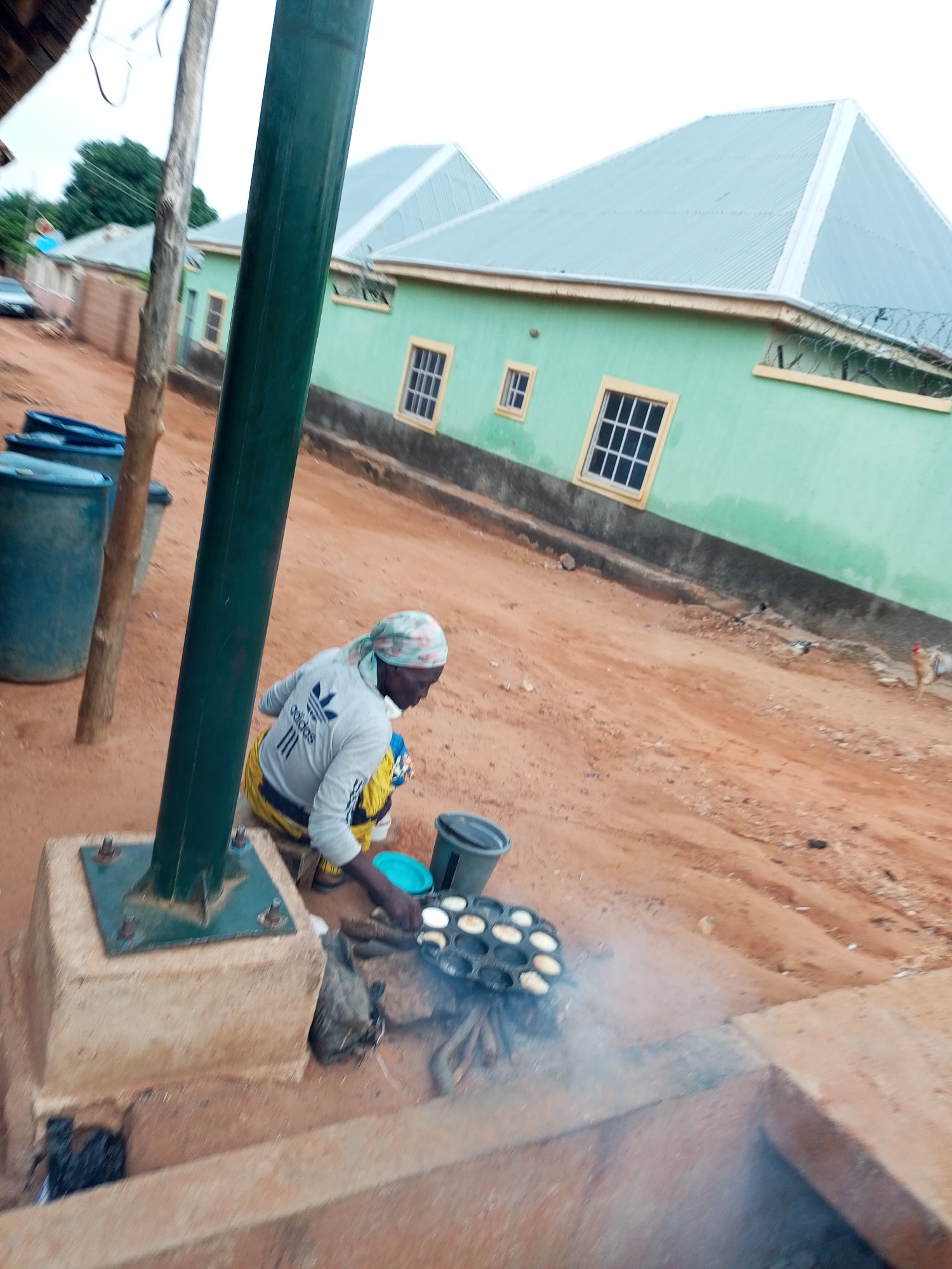 A person is cooking outdoors in a dirt area in front of a light green building with a gray metal roof. The person is wearing a light-colored headscarf and a gray long-sleeved shirt with the Adidas logo. They are kneeling and using a round pan with multiple small indentations, in which several small, white, circular food items are visible. A dark gray bucket is next to the pan, and a blue plastic bowl is on the ground near the person's hand. There are several large blue plastic barrels to the left of the scene, and a brown wooden pole is positioned vertically in the foreground. The ground is reddish-brown dirt, and there is some dry vegetation. In the background, there are additional buildings and trees, suggesting a residential or semi-rural area. The sky is overcast and pale. The general impression is of a casual, outdoor cooking setup.