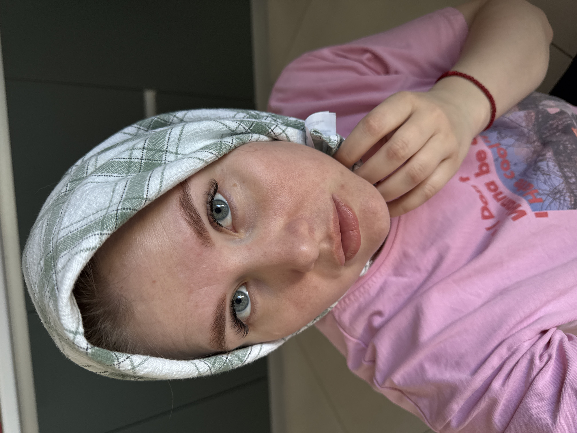 The image depicts a close-up, chest-up portrait of a female individual located indoors in Gornyi (Dvurechenskoe S/P), Russia. The subject has light skin, blue eyes with visible eyelashes, and light brown eyebrows. She is wearing a light green and white plaid headscarf, secured at the neck, and a pink short-sleeved t-shirt with a partial graphic print that includes the text "Don't Wanna be". A red beaded bracelet is visible on her left wrist. Her right hand is raised, with fingers gently touching her neck area, near where the headscarf is fastened. The individual's gaze is directed forward, towards the camera. The background is dark, uniformly out of focus, and includes a faint light-colored horizontal element in the upper left portion.