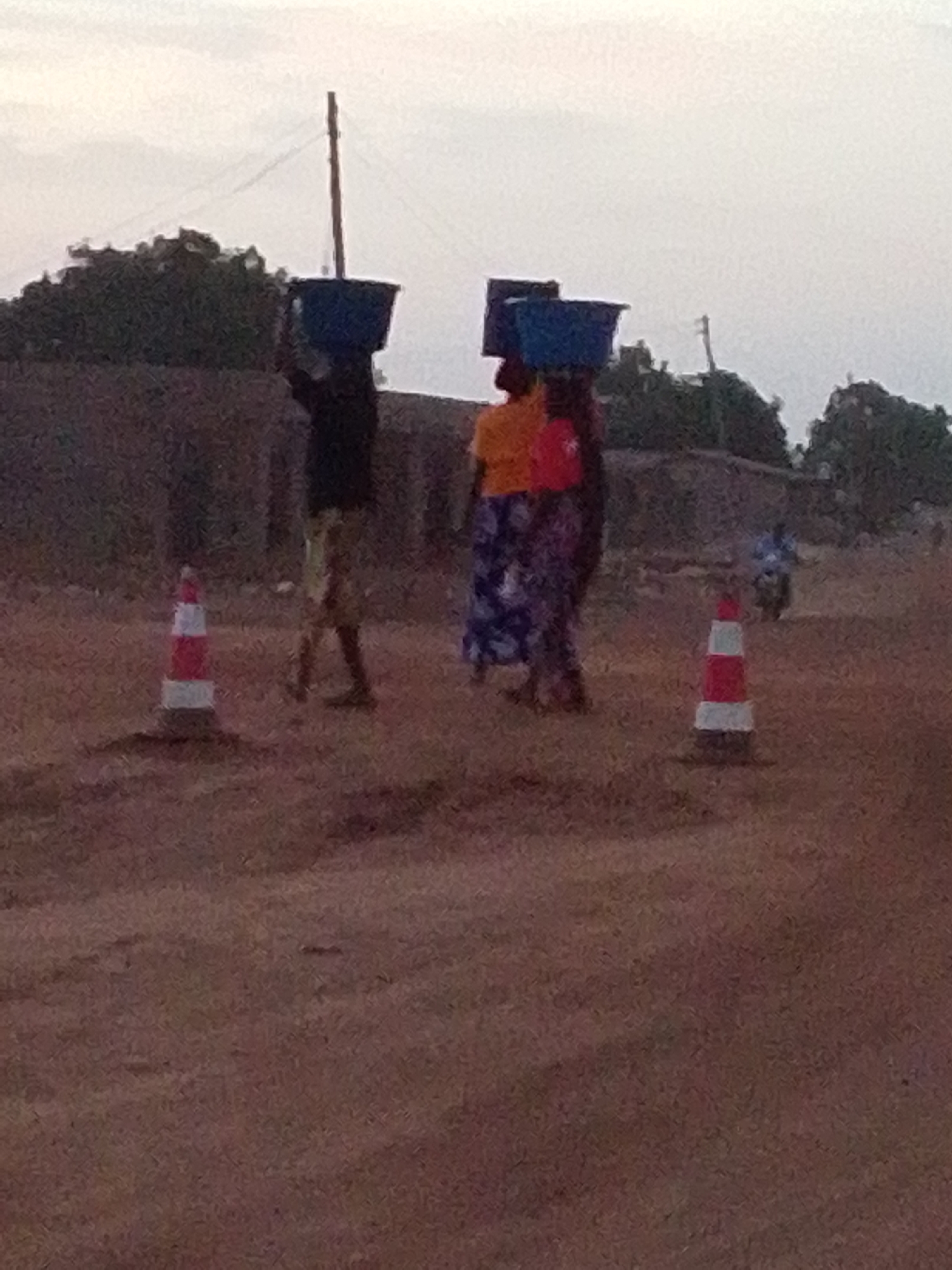 The image depicts a scene on a dirt road, likely during twilight or an overcast day. Three individuals are prominently featured, walking and carrying large, blue rectangular containers on their heads. This traditional method of transport suggests a rural or semi-urban setting, possibly in a warm climate.
In the foreground, the leftmost person wears a dark top and lighter-colored bottoms, balancing one blue container. To their right, two other individuals walk closely; one wears a bright orange or yellow top with a patterned skirt, also carrying a blue container. The third person is partially obscured by the second but also appears to be carrying a container. Their activity is consistent with daily chores or commerce.
Two red and white striped traffic cones are positioned on the ground, one on either side of the walking figures, marking an area or perhaps indicating a hazard. The ground itself is a reddish-brown, unpaved surface, appearing dusty. In the mid-ground, a tall, slender wooden pole stands upright, and behind the figures, there are low, indistinct buildings or walls and scattered trees. Further in the background, on the right side, a person on a motorbike or scooter is visible. The sky is light and hazy, indicating diffused light without direct sunlight. There is no visible text in the image.