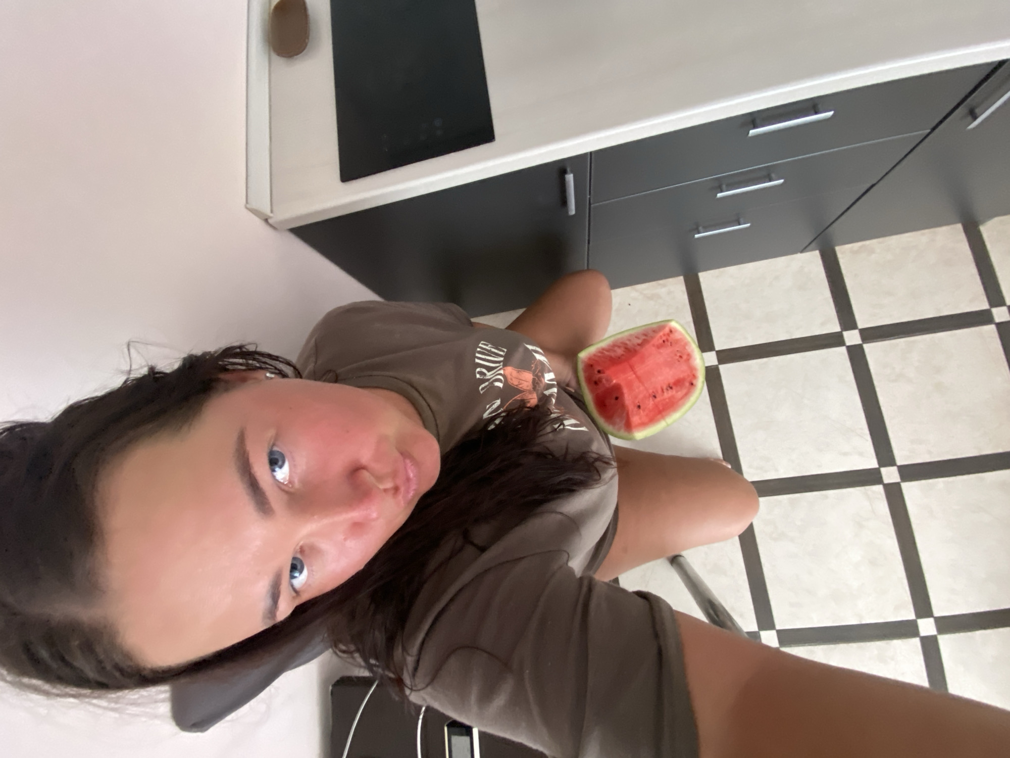 A young woman with dark hair and blue eyes is seated on a chair in a kitchen, holding a slice of watermelon. She is looking at the camera with her lips pursed. The kitchen has black and white tiled flooring and dark gray cabinets. The scene appears to be indoors, likely a residential kitchen. The city of Sochi, Russia, is not identifiable from the image.