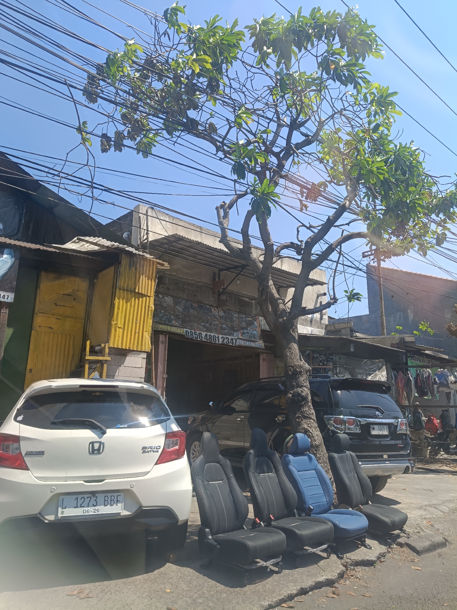 A sunny day in Surabaya, Indonesia, shows a street scene with parked cars, a workshop, and car seats displayed for sale. In the foreground, a white Honda Brio is parked, its rear facing the camera. To its right, several car seats, upholstered in black and blue, are arranged on the ground. Behind them, a black SUV is parked. A large tree with sparse green leaves grows in front of a weathered building that appears to be a workshop or a shop. The building has a corrugated yellow metal door and a sign that reads "JL MOBILSUK 0856 4801 234". Electrical wires crisscross overhead against a clear blue sky. The scene suggests a commercial area in Surabaya, possibly with a focus on automotive services.