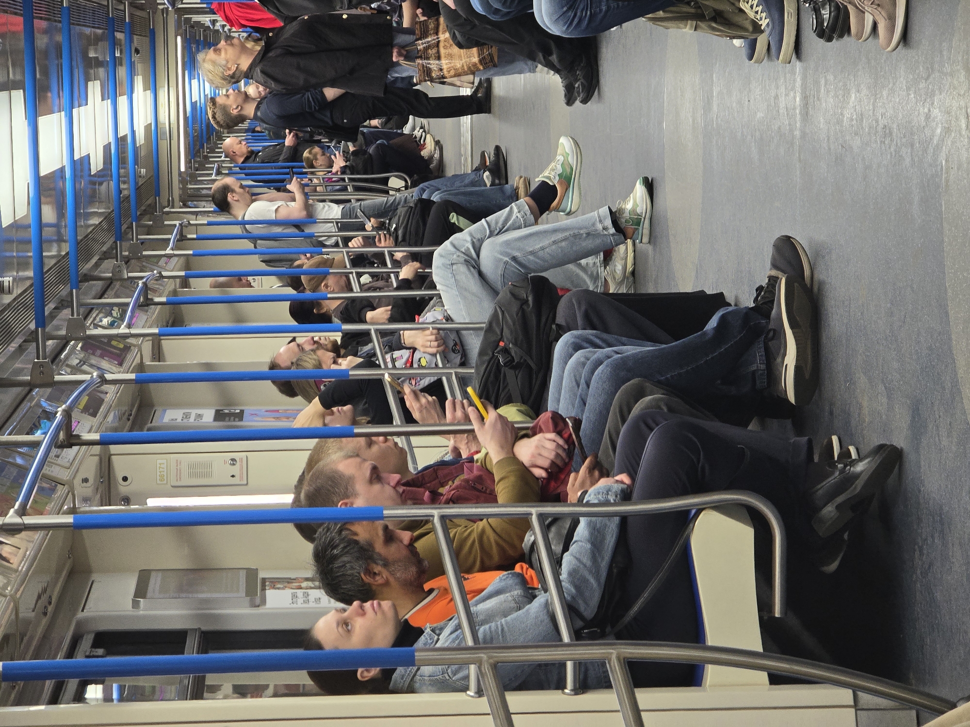 A low-angle, horizontally-oriented photograph shows the interior of a train car filled with passengers. The train car is brightly lit from above, with rows of overhead lights illuminating the scene. The interior of the train car is primarily neutral tones with accents of blue. The car is filled with rows of seating, oriented lengthwise. The passengers are seated and facing in the same direction, mostly looking forward. Several passengers are using smartphones. The subjects are dressed in a variety of casual attire, including jeans, t-shirts, and jackets. The floor of the train car is a neutral grey tone. The train is in motion. The location is Unknown, Unknown.