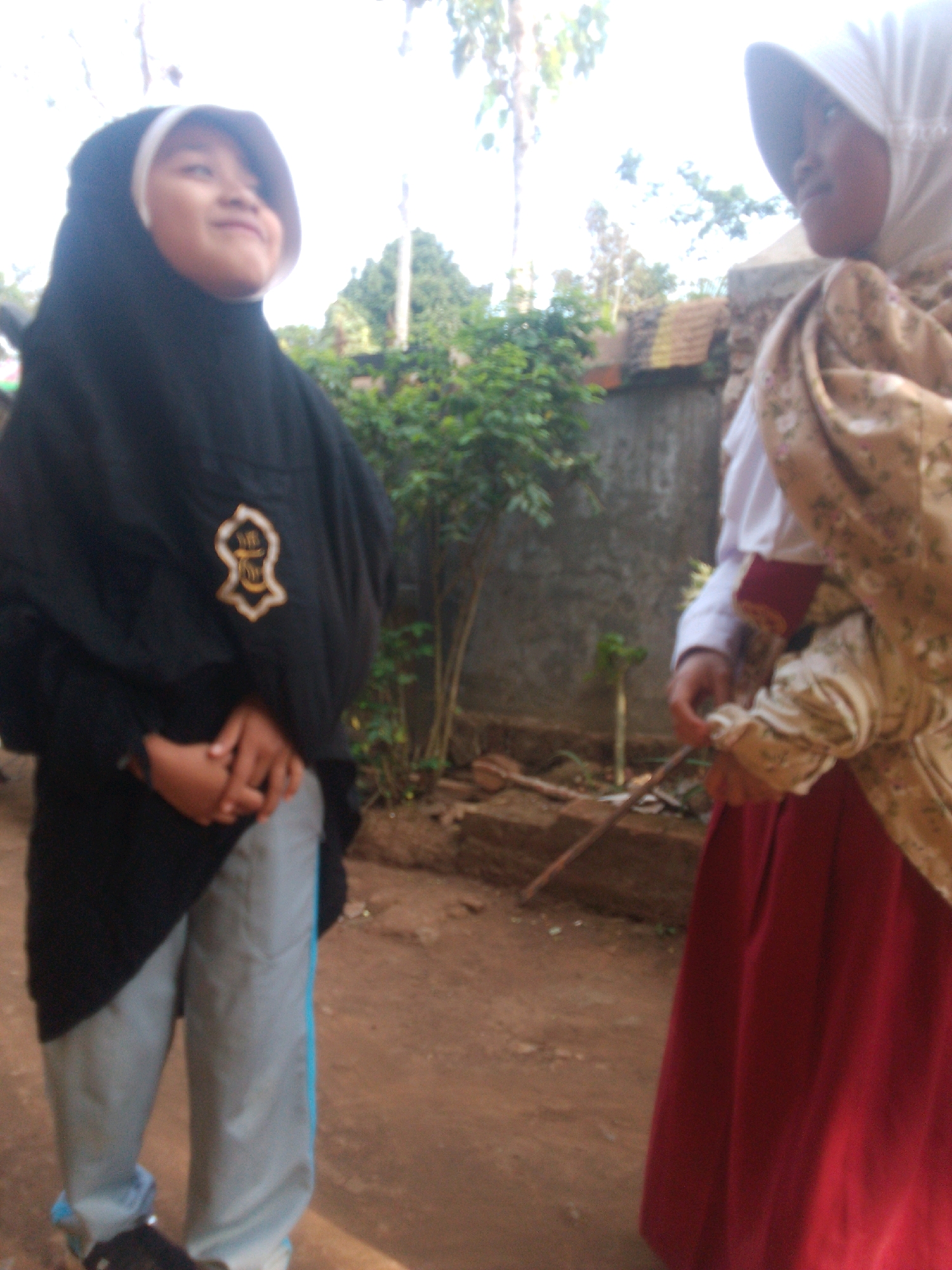 The image shows two young individuals outdoors, likely in the city of Betek, Indonesia. The person on the left is wearing a black hijab and light blue pants with a blue stripe. They are looking upwards and to the left. A gold emblem is visible on their clothing. The person on the right is wearing a light-colored hijab, a white shirt, and a dark red skirt. They have a patterned shawl draped over their shoulder. The background includes a concrete wall, trees, and some wooden debris. The ground is dirt.