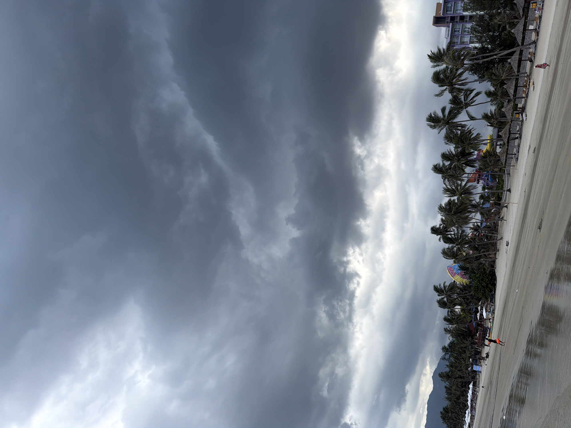A wide shot of a beach in Dong Ninh Hoa Ward, Vietnam, under a dramatic, overcast sky. The sand is wet and reflective, indicating recent rain or high tide. Palm trees line the edge of the beach, their fronds blowing in the wind. A few beach umbrellas and lounge chairs are visible, some occupied by a few people. In the background, a modern hotel with purple accents stands out. A person is walking on the wet sand in the foreground, and another is walking near the water's edge. The overall atmosphere is moody and turbulent, possibly due to an approaching storm.