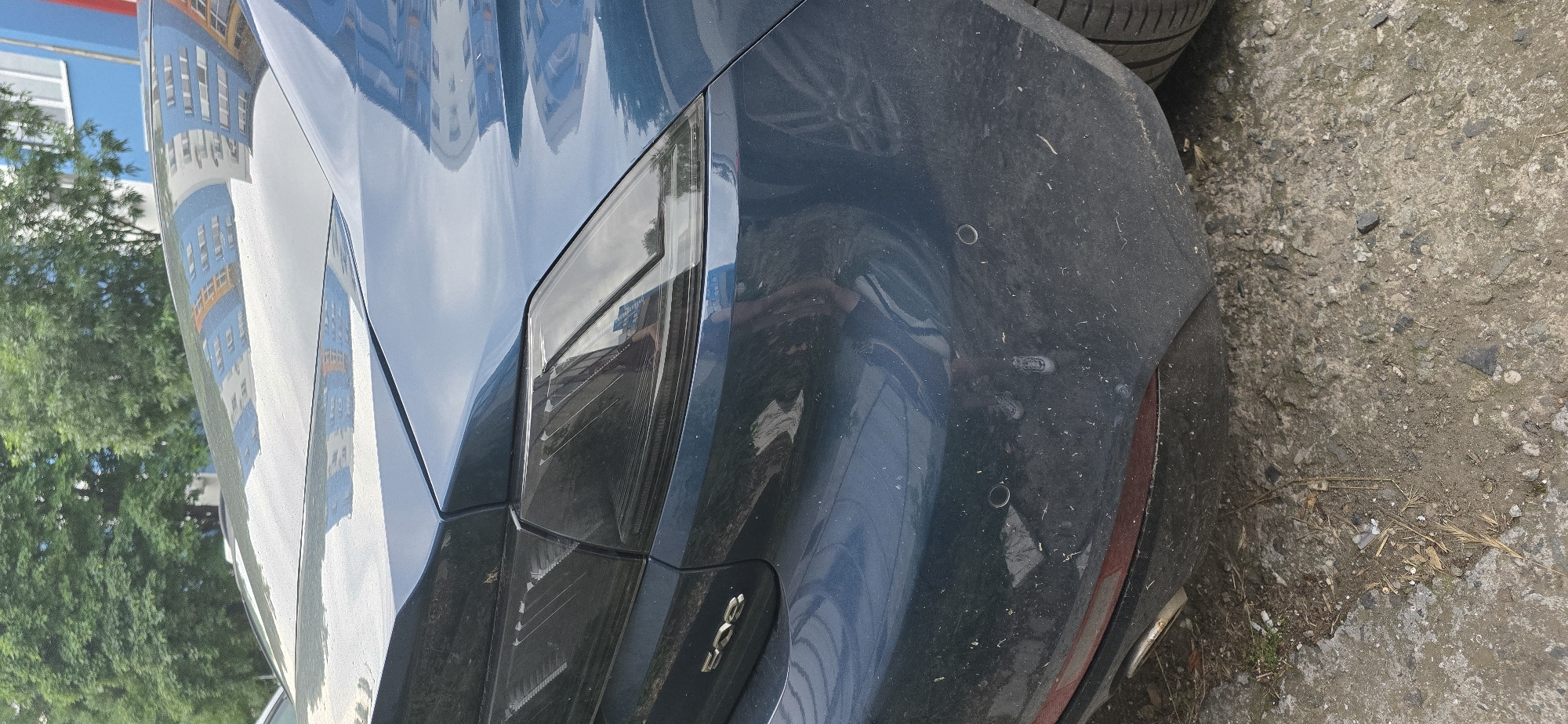 The image captures a close-up view of the rear quarter panel and bumper of a dark blue car, likely a Peugeot 308, parked outdoors on a sunny day in Burgas, Bulgaria. The car's metallic paint is highly reflective, prominently displaying the distorted reflection of a multi-story building featuring blue and white sections with numerous windows, alongside green foliage from trees.
The car's rear bumper is visibly dusty, with small particles and debris scattered across its dark surface. A part of the dark-tinted rear taillight assembly is visible, characterized by angular lines. On the bumper, at least two circular parking sensors are distinguishable. A chrome-colored "308" emblem is affixed to the trunk lid, clearly identifying the car model.
The vehicle is situated on a rough, grey ground surface that appears to be concrete or asphalt, littered with small stones, dirt, and some organic debris such as dried leaves and twigs. A black tire is partially visible in the upper and lower right sections of the frame, suggesting another vehicle or spare tire nearby. The bright reflections and clear visibility indicate daytime conditions. No people or specific activities are depicted.