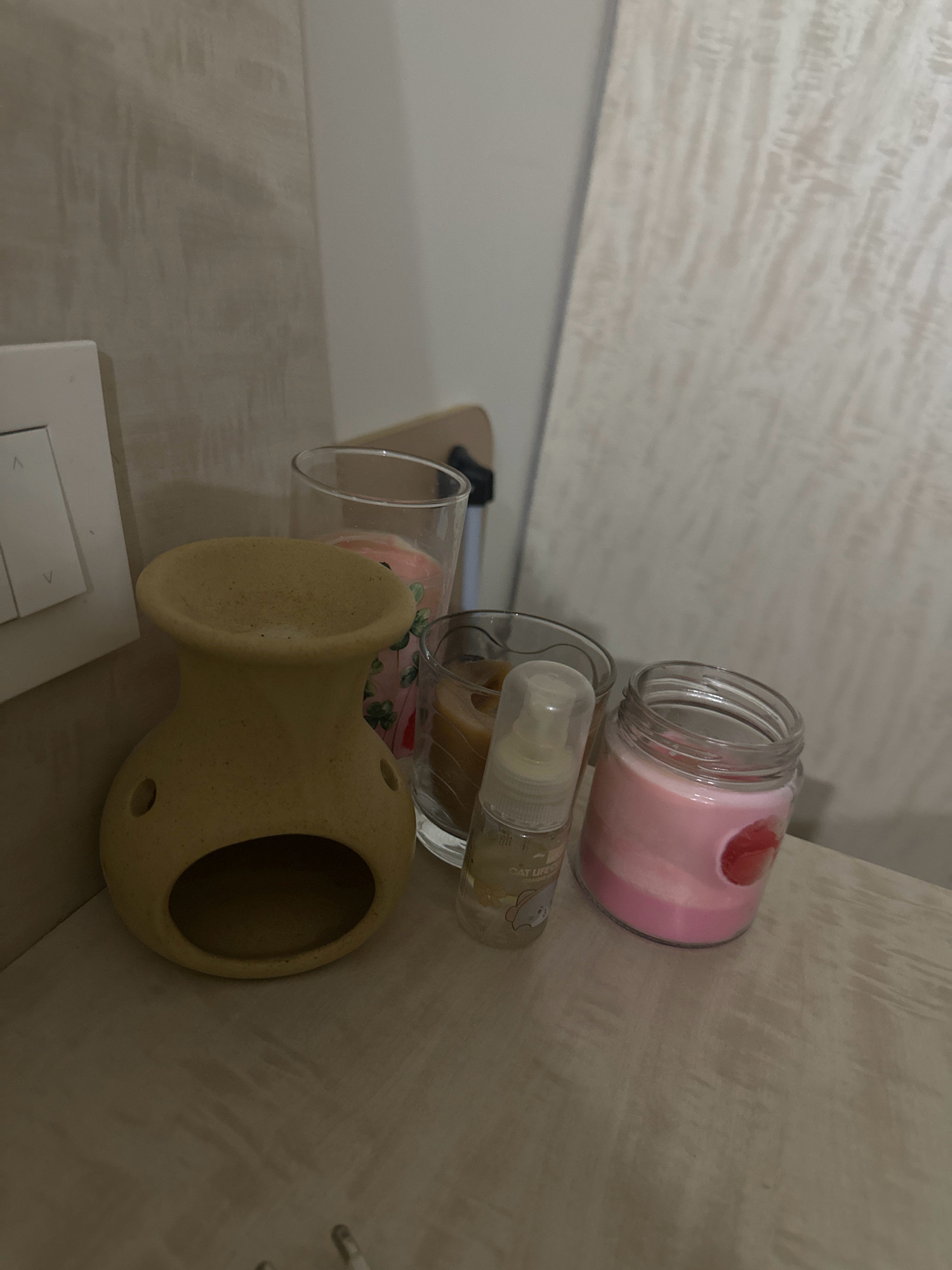 An indoor, eye-level, medium shot shows several decorative items on a wooden surface. On the left, a light yellow ceramic oil burner with an oval opening at the bottom and two circular holes on its sides sits prominently. To the right of the oil burner are three glass containers. The first, a clear glass, holds a pink candle with green clover patterns. Next to it is another clear glass containing a light brown candle. The smallest container, a clear glass bottle with a clear cap, stands in front of the other two. This bottle has a label with cartoon cat graphics and text. To the right of these is a clear glass jar filled with a pink and white layered candle. In the background, a white light switch is visible on a wall with a textured, light beige finish. A light-colored, textured panel leans against the wall on the far right. The scene is dimly lit, suggesting it is indoors and possibly nighttime. There are no people present.