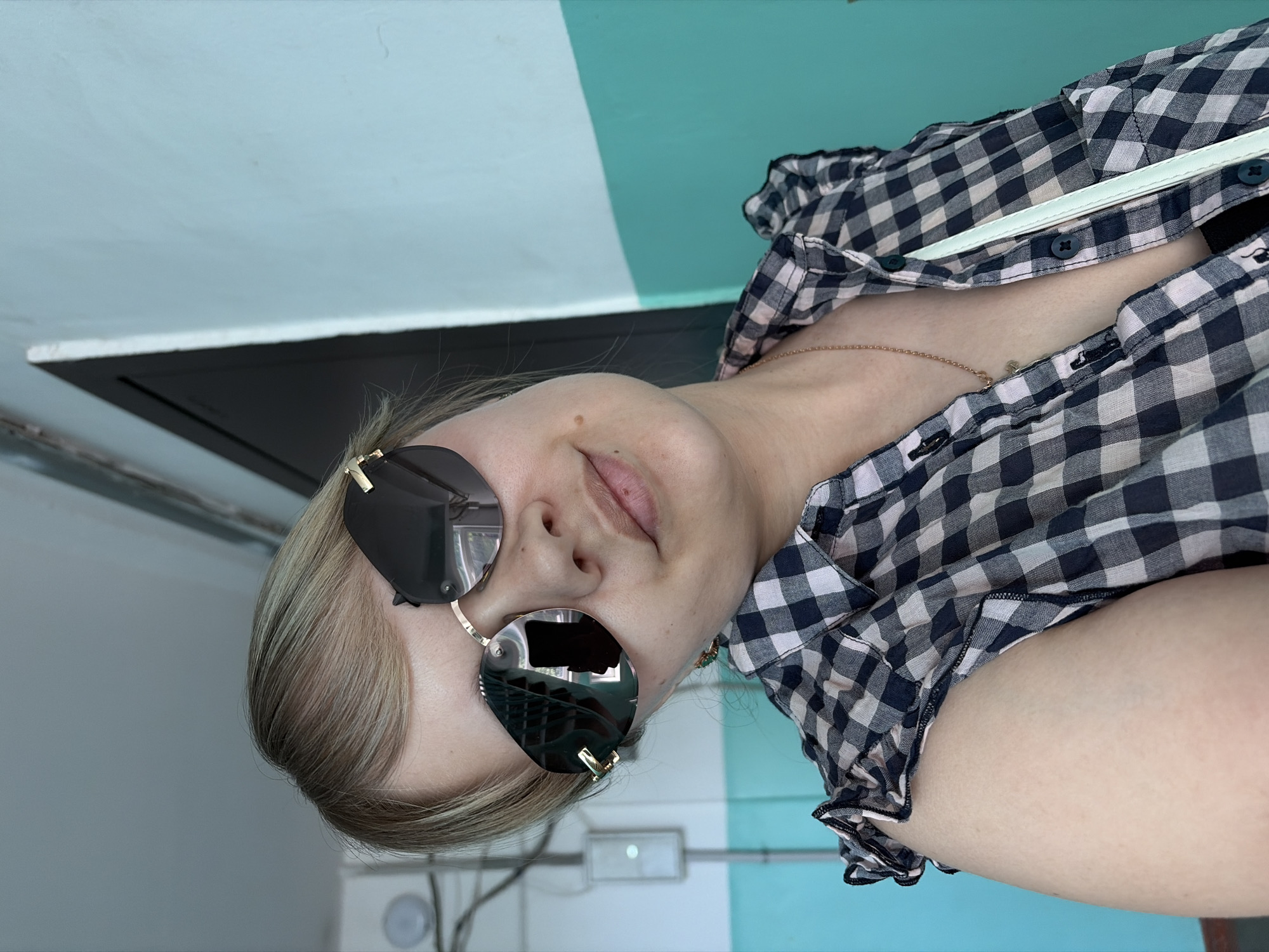 A woman wearing sunglasses and a checkered shirt is posing for a selfie indoors. The location appears to be in Chaikovsky, Russia. The background includes a wall painted with horizontal bands of white and turquoise. A black panel is visible in the upper left, and electrical outlets are visible lower down. The woman is wearing a thin gold chain around her neck.