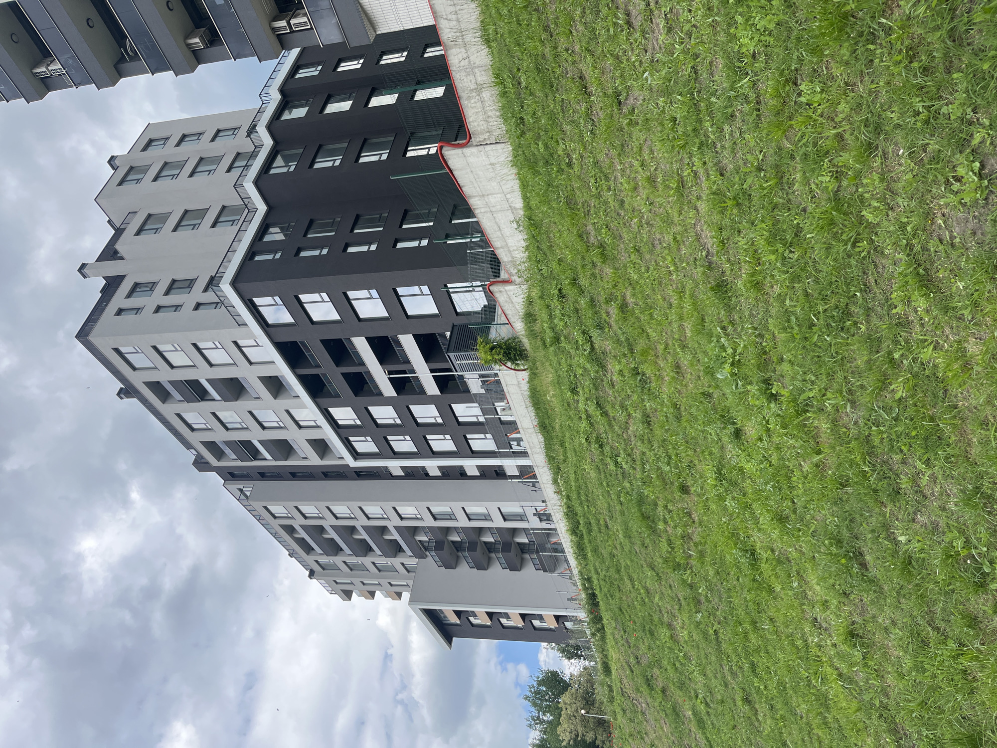 The image shows a modern apartment building in Varna, Bulgaria. The building has a two-tone facade, with grey on one side and black on the other. It has multiple stories and many windows. A patch of green grass is visible next to the building. The sky is cloudy. The building is viewed from a low angle. There are no people visible in the image.