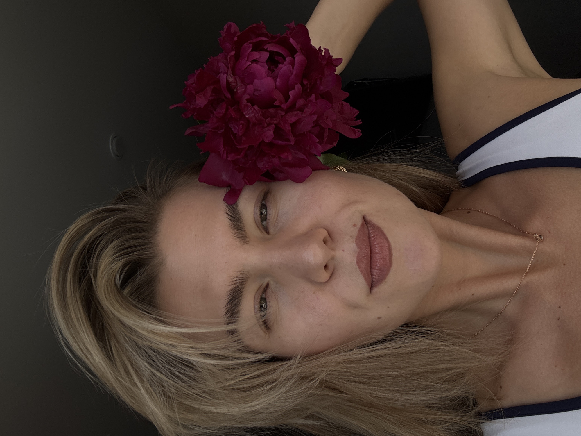 A head and upper torso portrait shows an adult female against a dark, featureless background, in the city of Unknown, Unknown. Her blonde hair is styled with volume, framing her face and falling onto her shoulders. She has fair skin, light-colored eyes, and dark, well-groomed eyebrows. Her lips are adorned with light-colored lipstick. A delicate, gold-colored chain necklace with a small pendant rests on her neck. She wears a white top with a dark-colored trim on the strap. A large, deep magenta-colored peony flower is held to the right side of her head, partially obscuring her ear and upper right cheek. Her right arm (viewer's left) is raised, indicating she is holding the flower. She gazes directly forward with a subtle, neutral expression. The background is uniformly dark, with a faint, blurry circular fixture visible in the upper right.