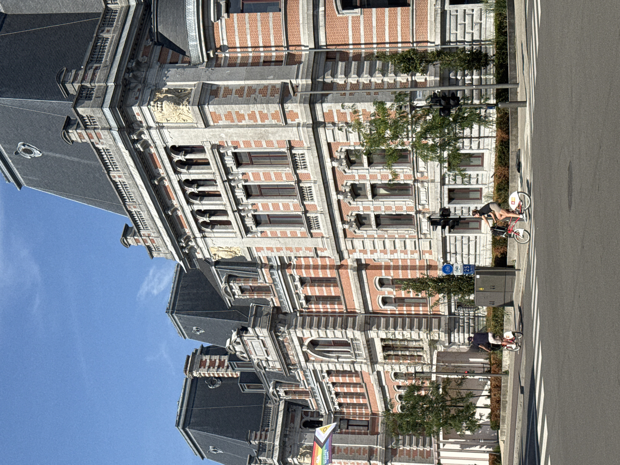 The image shows a sunny day with a clear blue sky above a large, ornate building with a red brick facade and grey stone accents, featuring a mansard roof. The building appears to be an official or historical structure. In front of the building, a street with a pedestrian crossing is visible. Two individuals are riding bicycles on the street. One cyclist is a person wearing a dark shirt and light-colored shorts, riding a bicycle with a white frame and red accents. The other cyclist is wearing a dark shirt and light-colored shorts and is riding a bicycle with a white frame. A traffic light is visible on a pole. A rainbow flag with a triangular insert in the upper left corner is attached to the building. The scene is located in the city of Antwerp, Belgium.