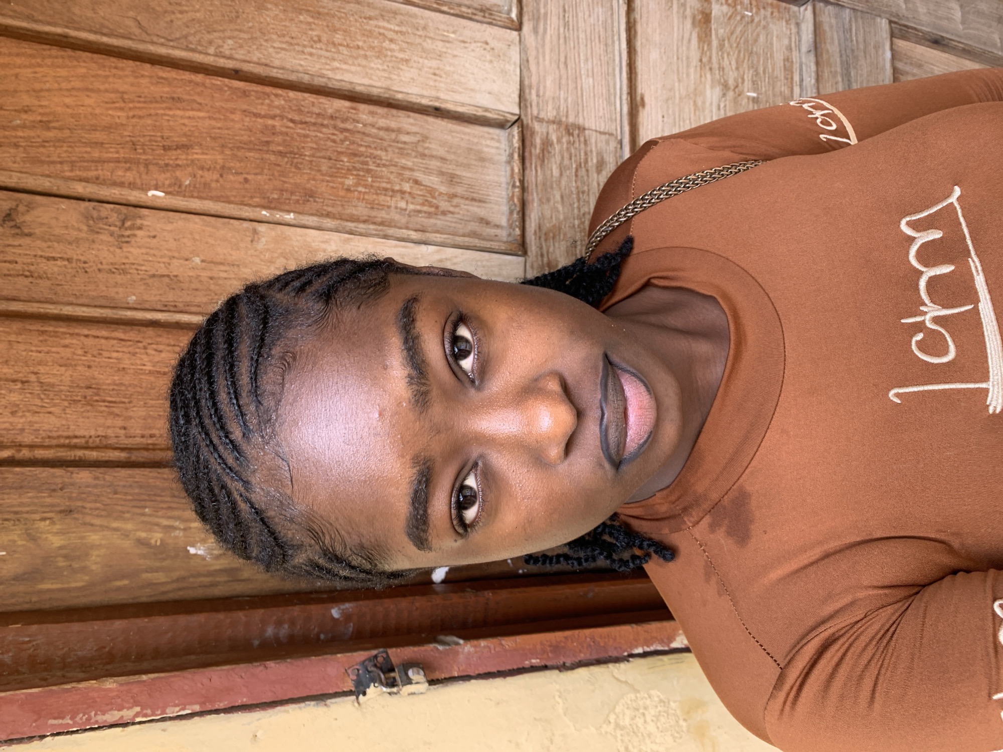 A young individual, appearing female, is depicted in a close-up portrait in Rafin Par, Nigeria. She has a dark skin tone and wears black cornrow braids with styled baby hairs along the hairline. Her dark eyes gaze directly forward, and her full lips are outlined in a dark liner with a lighter pinkish-nude fill. A noticeable sheen is present on her forehead and cheeks, suggesting perspiration or an oily complexion.
She wears a long-sleeved, crew-neck shirt in a rusty brown or terracotta color. The shirt features white or cream-colored cursive embroidery on the chest, partially reading "I am," with additional script visible on the right sleeve. A gold-toned chain strap, likely from a bag, is visible over her right shoulder.
The background directly behind her consists of vertical wooden planks, suggesting a door or wall panel, characterized by a light-to-medium brown wood grain. To the left of the subject, a section of a peeling yellow wall or pillar is visible, revealing an underlying reddish-brown layer. A metal padlock and hasp are attached to a vertical reddish-brown support at the junction of the yellow wall and the wooden background element. The scene is illuminated by soft, even light.