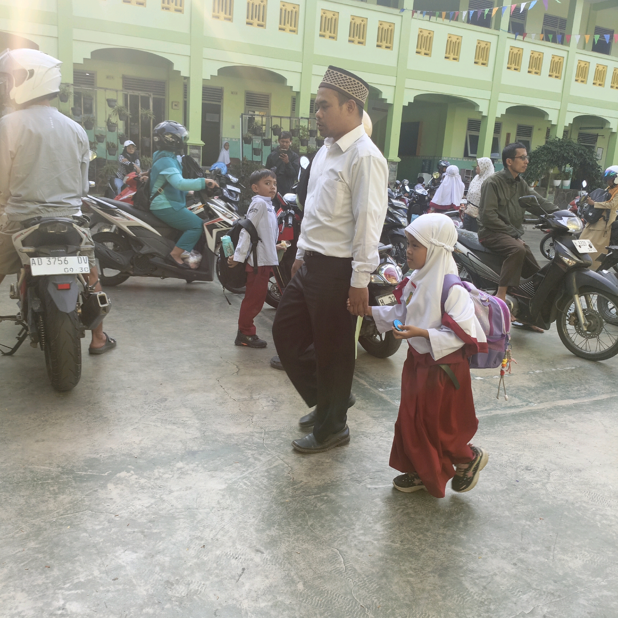 The image depicts a scene outside a light green building, likely a school, in Salatiga, Indonesia. The building has yellow-framed windows and colorful triangular flags strung across the upper level. The ground is paved with concrete, showing some cracks and light smudges.
In the foreground, a young girl wearing a white hijab and a red skirt, with a purple backpack, is walking to the right. She is holding a small blue object in her right hand. To her left, a man in a white long-sleeved shirt and black pants, wearing a patterned peci (a cap common in Indonesia), is walking in the same direction. He is holding the girl's left hand. Behind them, a young boy in a white long-sleeved shirt and red pants, with a backpack, is walking towards the left.
Several motorcycles are parked in the middle ground and background. One motorcycle in the far left has a license plate reading "AD 3756 DV 09.30". A person wearing a white helmet is seated on this motorcycle. Another person in a turquoise outfit and black helmet is riding a motorcycle behind them, to the left of the man in the white shirt. Further back, a person in a dark green jacket is riding a motorcycle. Other people, including those in hijabs, are visible near the motorcycles. The overall atmosphere suggests a busy morning, possibly the start or end of a school day.
