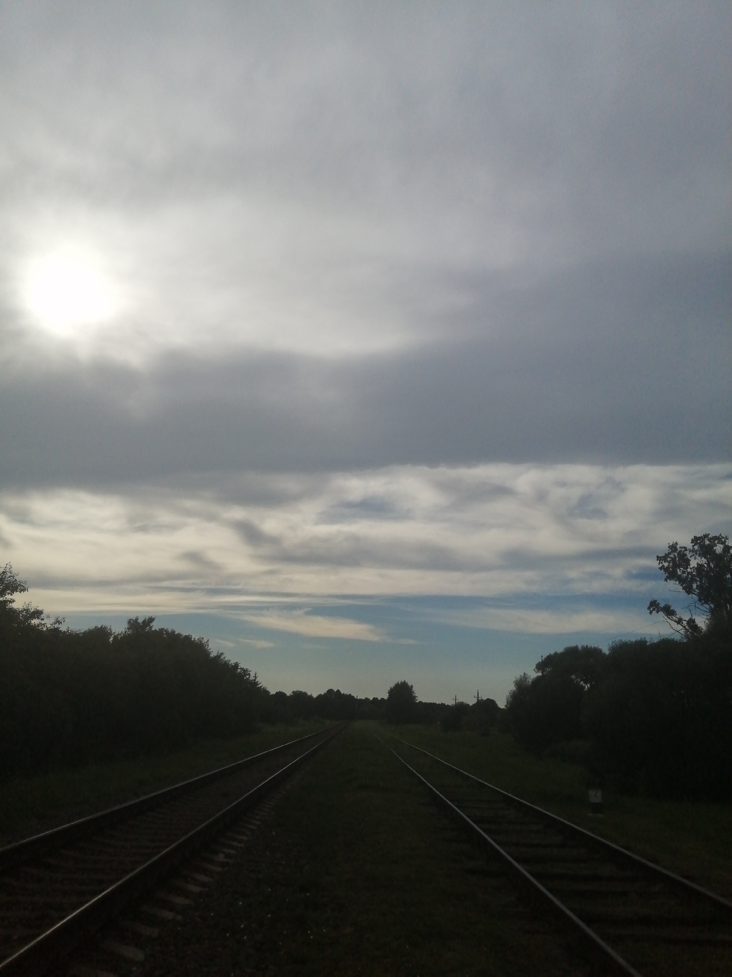 The image shows a railway track stretching into the distance under a cloudy sky. The sun is partially visible through the clouds on the left side of the frame. Dense foliage, likely trees, lines both sides of the railway track. The scene appears to be in a rural or semi-rural area. The location is not discernible from the image itself; no landmarks or specific features indicate it is in the city of Slonim, Belarus. There are no people or visible actions taking place.