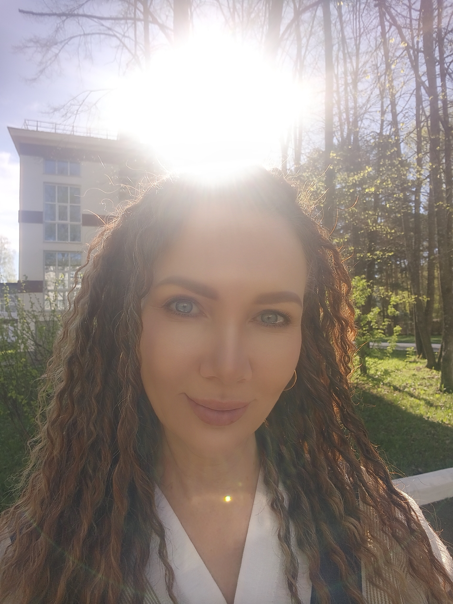 A person with long, curly brown hair, wearing a white collared shirt, is taking a selfie outdoors. The subject has light eyes and a slight smile. The background is bright, with a building to the left and trees to the right, heavily obscured by sunlight. The location appears to be in a park or near a wooded area, in the city of Unknown, Unknown.