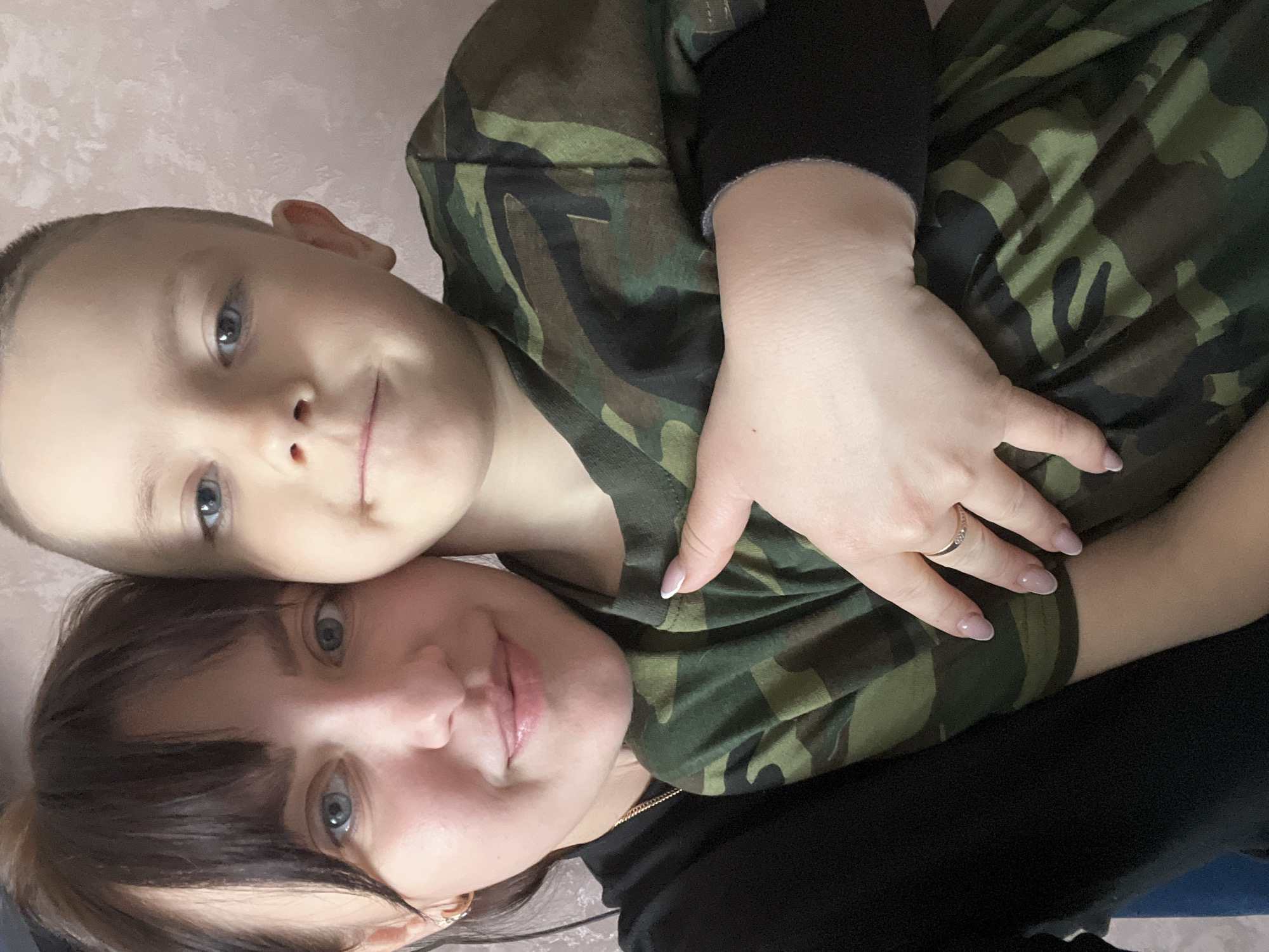 A young boy and a woman, presumably mother and son, are posing for a selfie. The boy is wearing a camouflage patterned shirt and the woman is wearing a black shirt. The woman has her arm around the boy. The background is a light pink color. The location is Марковский, Russia.