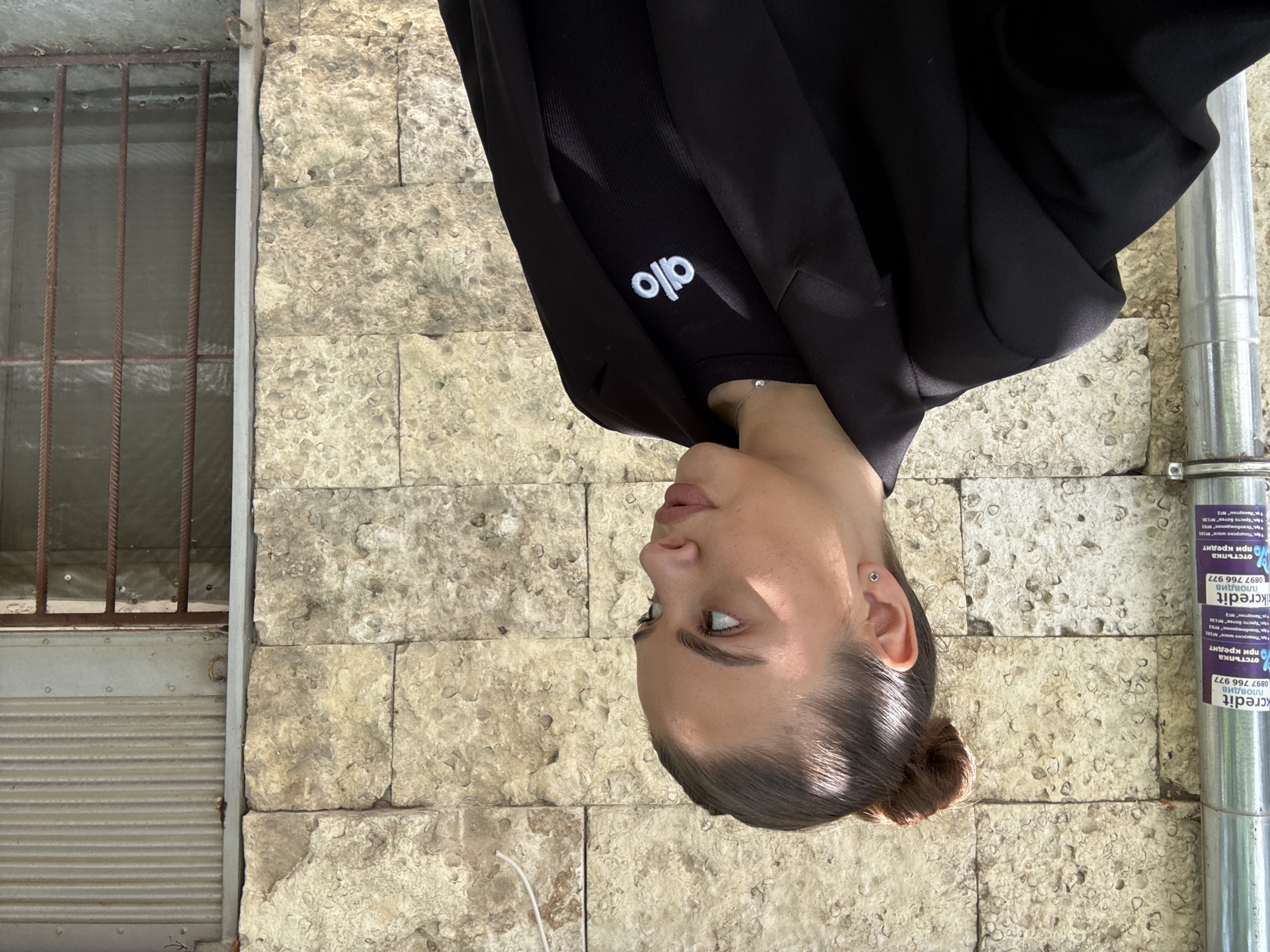 The image shows a woman wearing a black sweatshirt with the "Alo" logo on the sleeve. She is standing outside, next to a stone wall with a barred window. The wall also has a metal pipe attached to it, with "kcredit" printed on it in Bulgarian. The location is likely in Plovdiv, Bulgaria, based on the Bulgarian text on the pipe. The image is rotated 180 degrees.