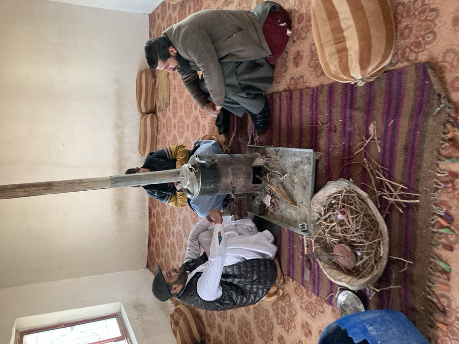 Inside a light-colored room in Sanjawi, Pakistan, three adult males are seated on a patterned red carpet. A cylindrical black metal wood-burning stove with a tall flue pipe dominates the center foreground, resting on a metal tray containing wood fragments. A kettle or teapot sits on top of the stove. To the left, a man with a dark beard, wearing a white shalwar kameez, dark quilted vest, and a dark cap, sits on the floor looking towards the right. Partially obscured behind the stove and pipe, a second man wears a dark jacket with yellow sleeves and light blue shalwar kameez, appearing to attend to the stove. To the right, a third man with a prominent dark beard, dressed in a light brown jacket and dark grey shalwar kameez, sits on a red cushion, looking towards the man on the left. In the immediate foreground, an oval-shaped basket containing small sticks and kindling rests on a striped maroon and brown rug with fringes, which also underlies the stove. Loose sticks are scattered around the basket. A partial blue plastic container and a metal dish are visible in the lower-left corner. Several striped and plain cushions are arranged against the walls and on the floor. A red-framed window is visible on the left wall. The men appear to be engaged in conversation while gathered around the heat source.