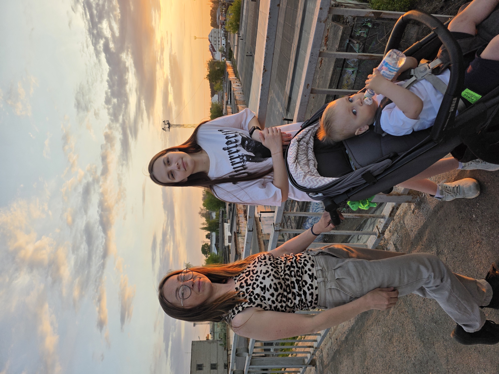 The photograph shows two young women and a baby in a stroller on an elevated walkway or bridge in the city of Chaikovsky, Russia.
One woman is wearing a white t-shirt and has long brown hair. She is standing near the stroller. The second woman is wearing a leopard print top and light-colored trousers. She also has brown hair and is wearing glasses. They are standing on what appears to be a pedestrian bridge or walkway, with industrial buildings visible in the background.
A baby is in the stroller, drinking from a bottle. The setting is sunset, as indicated by the orange and pink hues in the sky and the lighting on the subjects. The overall mood is casual and peaceful, capturing an everyday moment in the city. The architecture in the background and the lettering on the bottle are consistent with a location in Russia.