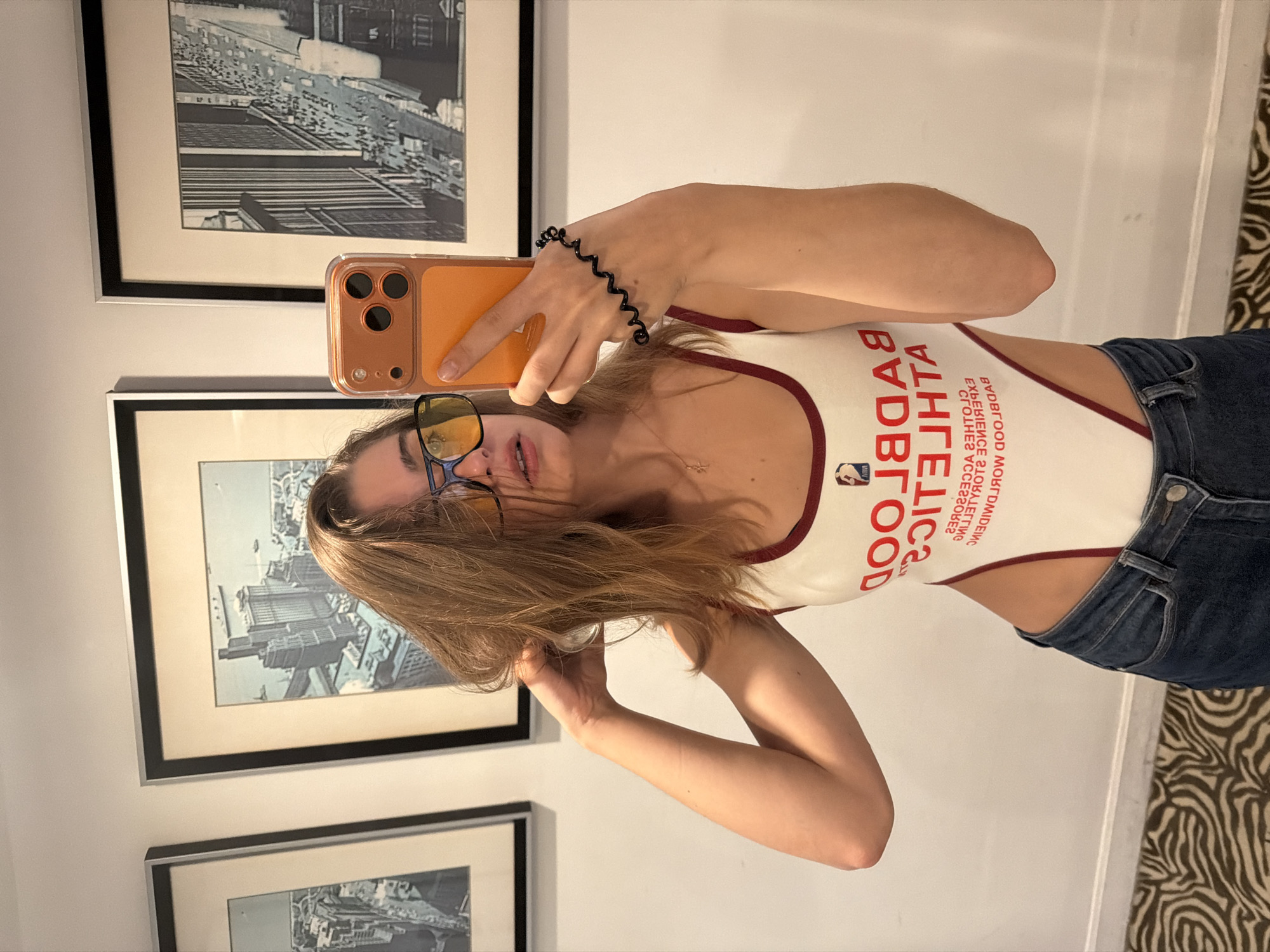 A young woman with blonde hair, wearing blue-tinted yellow sunglasses, a white bodysuit with red trim and text reading "BADBLOOD ATHLETICS", and blue jeans, stands in front of a white wall. She is holding an orange cell phone in her right hand, taking a selfie. There are three framed pictures on the wall behind her. The picture on the left is a black and white aerial view of a city with buildings and a ship. The middle picture is also a black and white aerial view of a city with buildings and a body of water. The picture on the right is a black and white cityscape with buildings and people. The floor is covered with a zebra print rug. The image does not provide any information or context to indicate the location is in Hong Kong, China.