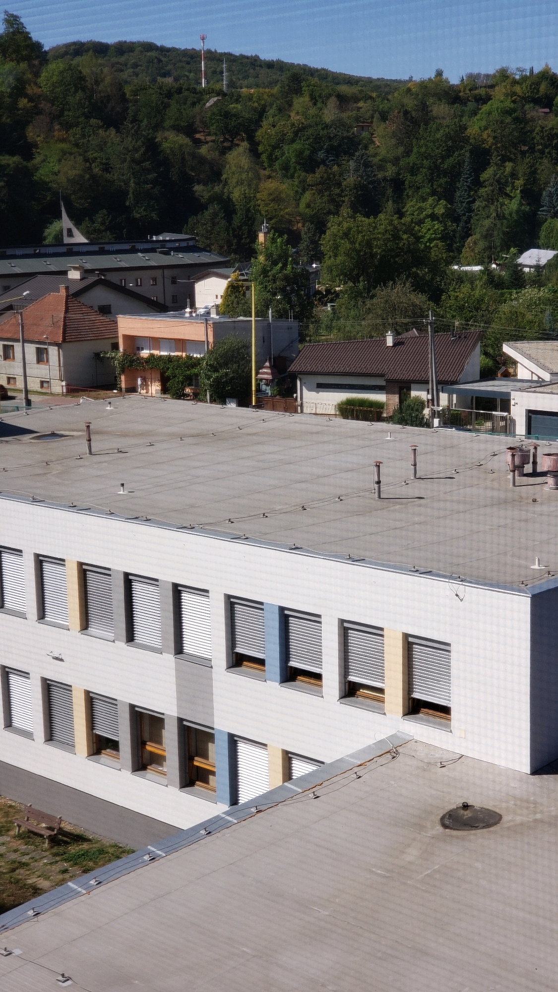 This is an aerial, daytime photograph taken from an elevated perspective, looking down on a modern building with a flat, grey roof. The building is multi-story, with a white facade and numerous windows. These windows are covered with metallic, slatted blinds that are mostly closed. Some sections of the facade are accented with vertical bands of color: pale yellow, light blue, and grey. The building is located in Prešov, Slovakia, evidenced by the architectural style and the surrounding greenery which suggests a temperate climate.
To the left of the main building, a lower, flat-roofed structure is visible, with a concrete surface and several ventilation pipes and electrical conduits. Further in the background, several other buildings with pitched roofs, some residential and some appearing to be school or institutional buildings, are nestled amongst dense green trees. The trees form a thick, undulating forest that covers a hillside. A telecommunications tower with red and white markings is prominently visible on the crest of the hill.
In the foreground, the lower portion of the photograph reveals a paved walkway and a small grassy area with a wooden park bench. The overall scene depicts an urban or suburban environment characterized by a mix of modern architecture and natural landscape. The sky is clear and blue, indicating good weather.