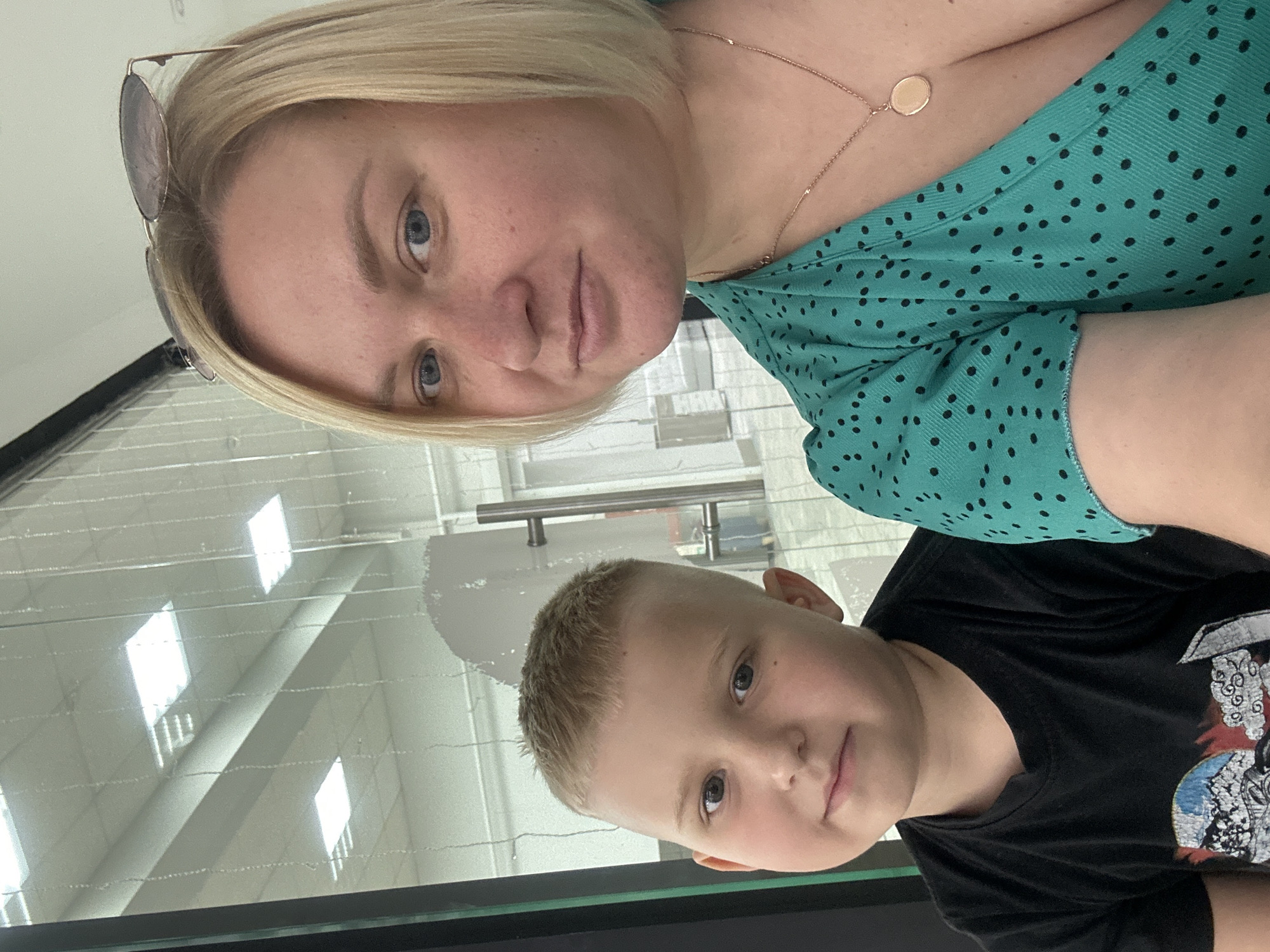 The image is a selfie taken indoors, possibly in a commercial establishment, with a glass partition reflecting lighting fixtures on the ceiling. On the left side of the frame, a young boy with short, light-colored hair and gray eyes is looking forward. He is wearing a black t-shirt with a graphic print. To his right, a woman with blonde bobbed hair, blue eyes, and light skin is also looking towards the camera. She is wearing a teal dress with black polka dots and a delicate gold necklace with a circular pendant. Sunglasses are perched on top of her head. The background through the glass appears to be a hallway or commercial space with multiple illuminated ceiling panels. There are no discernible elements in the image that would place the scene specifically in Chaikovsky, Russia.