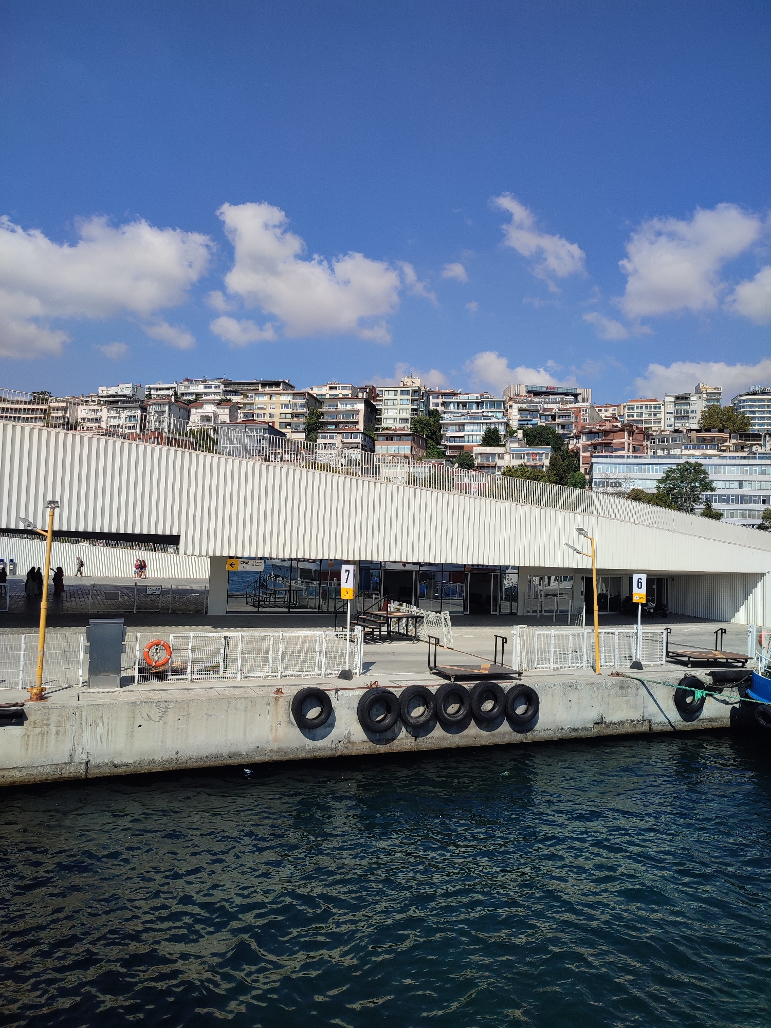 The image shows a waterfront scene with a modern ferry terminal building in the foreground, situated in the Beyoğlu district of Istanbul, Turkey. The terminal has a distinctive white, vertically slatted facade. A concrete pier runs along the front of the building, with several black tires acting as fenders. The water in the foreground is dark blue and choppy.
Behind the terminal, a hillside is densely populated with buildings of varying architectural styles and colors, indicating an urban environment. The sky is a clear blue with scattered white clouds.
On the pier, there are several people walking. Some are near the entrance of the terminal, while others are further down the pier. There are yellow signs with numbers and text, including one with "7" and another with "6", likely indicating ferry platforms or gates. A bright orange life preserver ring is visible on the pier. Some metal structures and carts are also present on the pier. A yellow lamppost stands on the left side of the image. A portion of a blue boat is visible on the right edge of the frame.