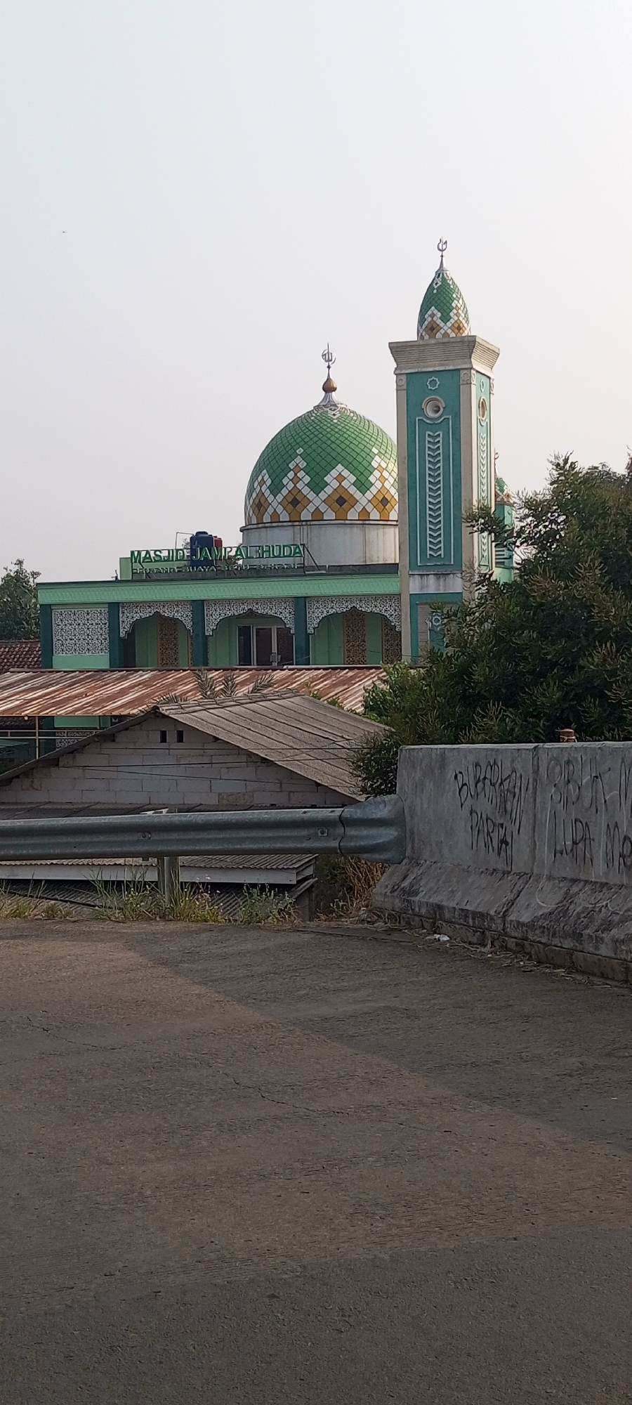 The image shows a mosque with a green dome and a minaret, located in Lubangbuaya, Indonesia. The mosque is named "MASJID JAMPAL BUDA". It is a sunny day with a clear sky. In the foreground, there is a road and a concrete barrier with graffiti on it. In the background, there are some houses with rusty roofs and trees. The overall scene depicts a typical urban landscape in Indonesia.