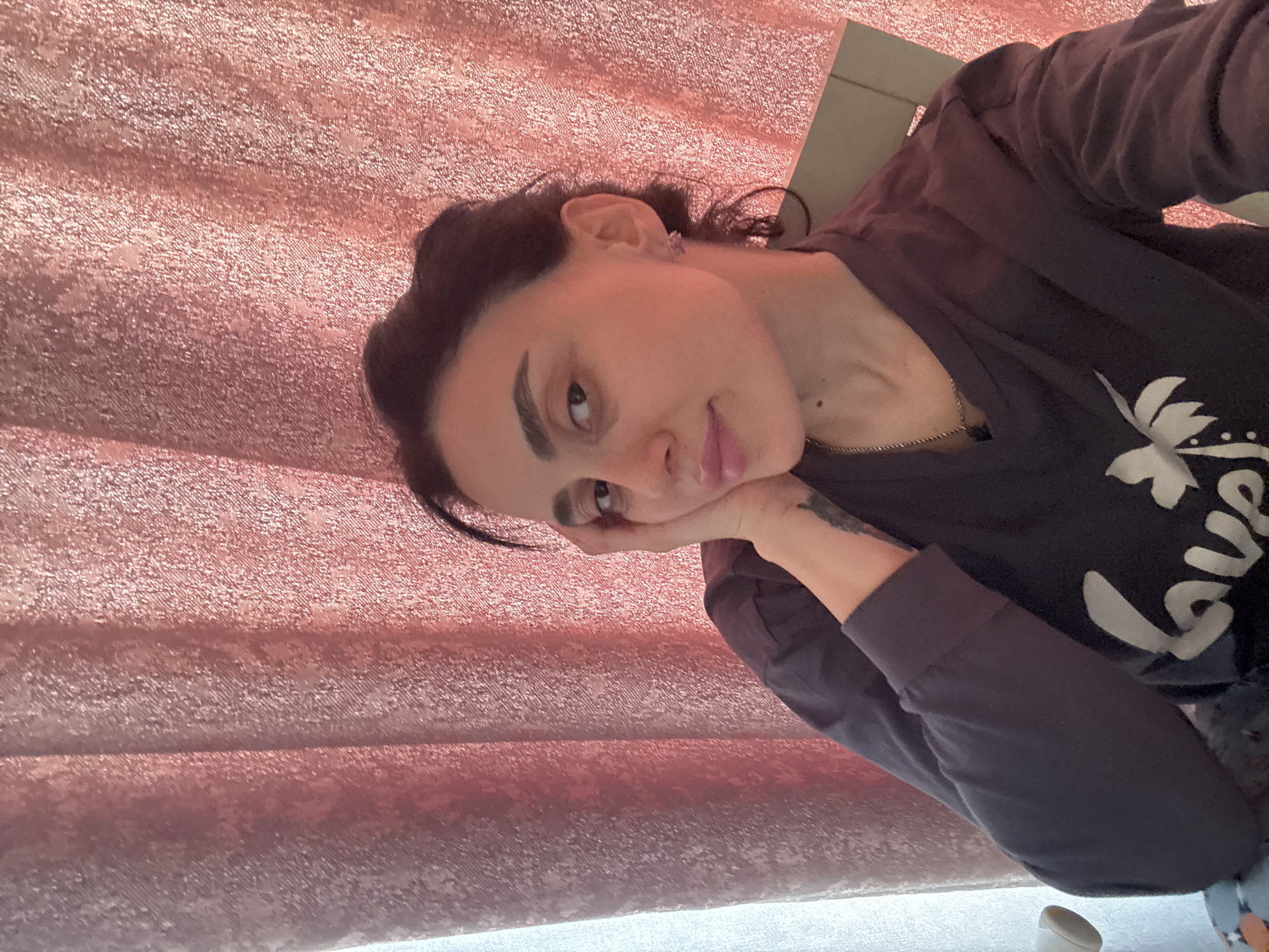 A medium indoor portrait features a woman with dark hair and fair complexion, shown from the chest up, leaning her head on her right hand. She looks towards the viewer with a neutral, slightly pensive expression. Her dark hair is styled back, highlighting her well-defined eyebrows and lips, which are colored with light pink gloss. She wears a dark, long-sleeved top, possibly charcoal grey or dark blue, adorned with a white graphic design. Partial text "L ve" (likely "Love" or "Lover") is visible on the shirt, alongside a floral element. A delicate gold chain necklace is visible, and she has a small earring in her right ear. A tattoo is subtly visible on her left forearm. The background is dominated by a shimmering, textured rose gold or pink fabric, appearing to be heavy curtains, providing a soft, warm ambiance. The image is captured at a 90-degree rotation. Although the location context is Tbilisi, Georgia, there are no specific visual cues within the image itself to confirm this. The lighting is soft and even, typical of an indoor setting.