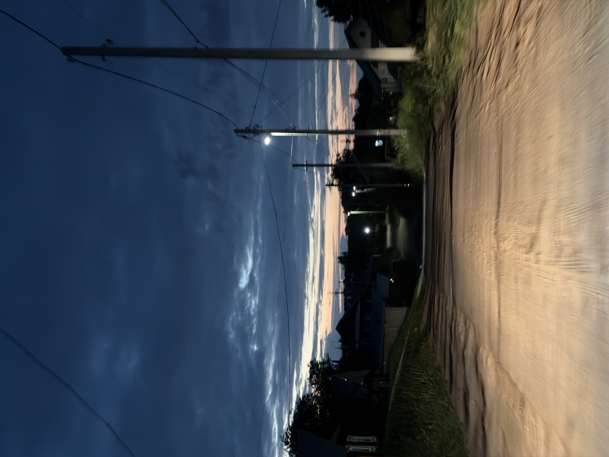 The image depicts a rural dirt road at dusk in the city of Fokinsky, Russia. The road is illuminated by the headlights of an unseen vehicle, casting a warm glow on the textured surface. On either side of the road, there are grassy verges and shadows cast by overhead power lines and poles. In the background, silhouetted houses and trees are visible under a deep blue, cloudy sky streaked with hints of orange and pink from the setting sun. Several streetlights are also illuminated along the road, providing additional points of light in the deepening twilight. The scene suggests a quiet, possibly isolated, evening in a small town.