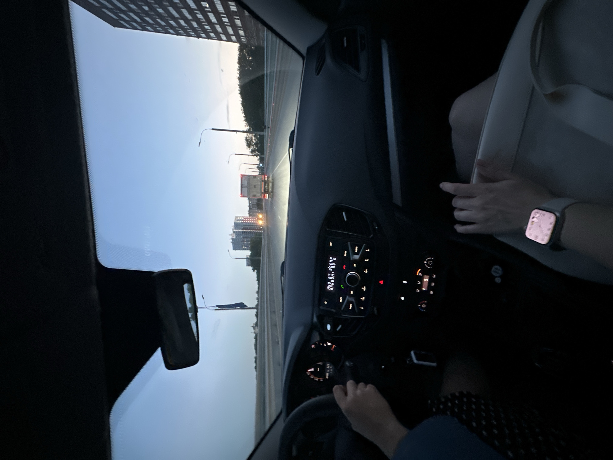 A medium shot depicts the interior of a car driving along a road in Perm, Russia, at twilight. The driver's hands are visible on the steering wheel. A passenger's hand, wearing a smartwatch, rests on the passenger seat. The dashboard and instrument panel are illuminated. Outside the windshield, streetlights line the road, and buildings can be seen in the distance. A large apartment building is also visible to the left. A bus can be seen driving along the road.