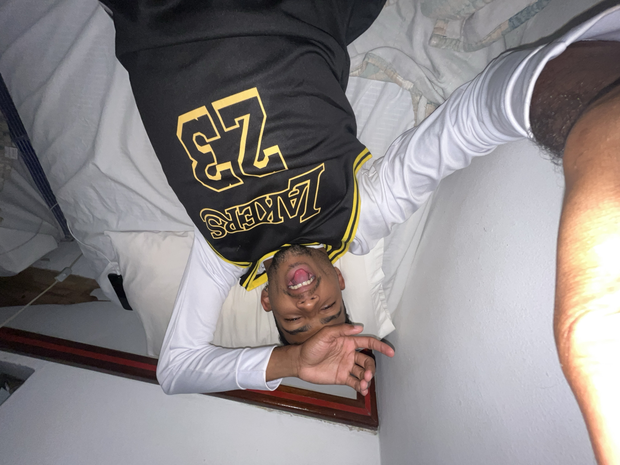 A dark-skinned man lies on his back on a bed. He is wearing a black and yellow Lakers jersey with the number 23 and a white long-sleeve shirt. He is holding his right hand near his face and sticking out his tongue. He appears to be taking a selfie. The bed has white sheets and pillows. The room has white walls. A portion of a dark wood frame is visible in the lower-left corner. The location is an interior space, possibly in Porlamar, Venezuela.
