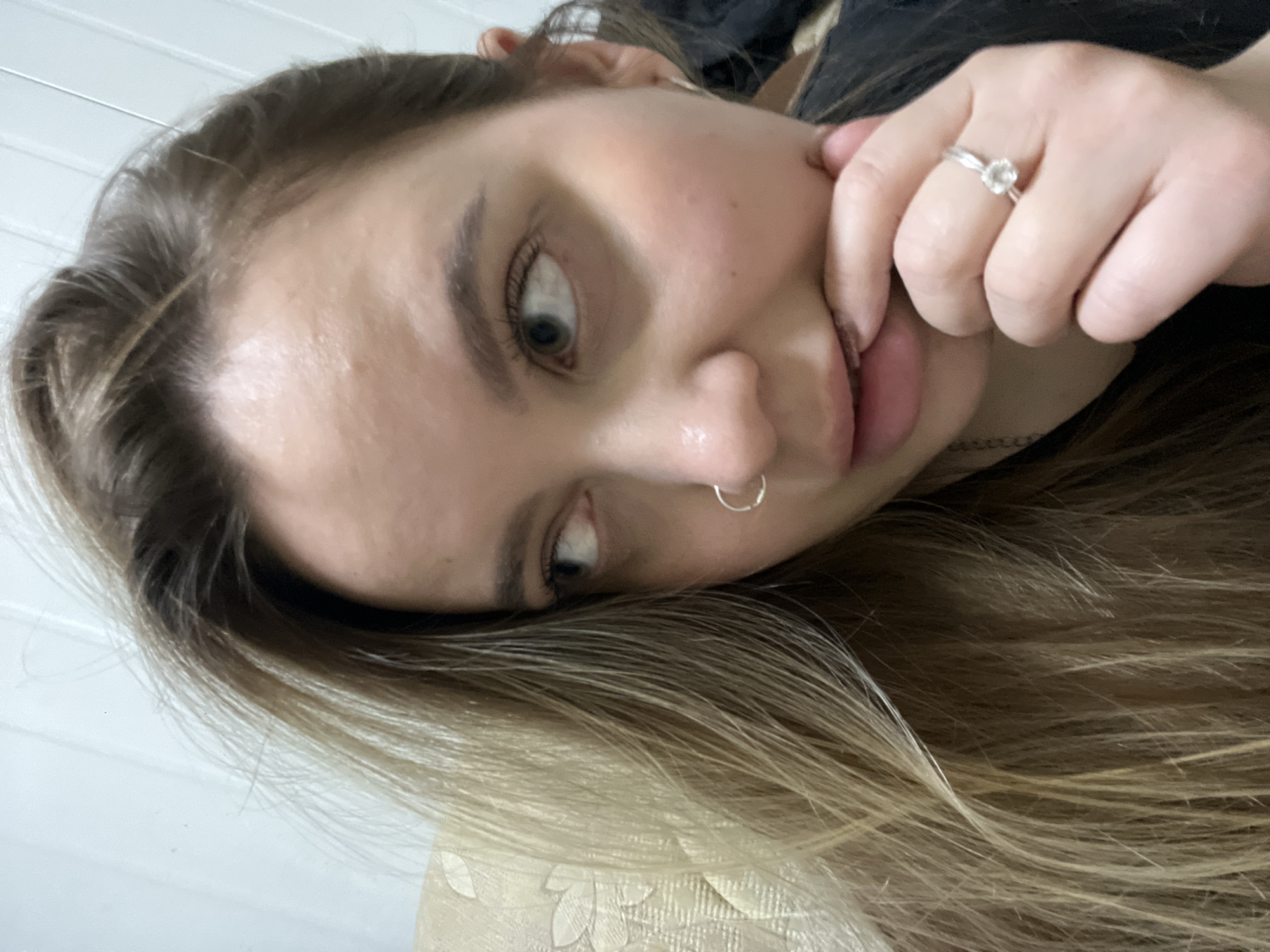 A close-up, tilted selfie shows a young woman with light brown and blonde hair. She has fair skin, blue eyes, and a silver hoop nose ring. Her hand, adorned with a silver ring with a floral design, is near her face. She appears to be resting her head on a light-colored, patterned surface. The background includes a glimpse of white vertical paneling. No information is available to definitively place the scene in Novopokrovskaya, Russia.