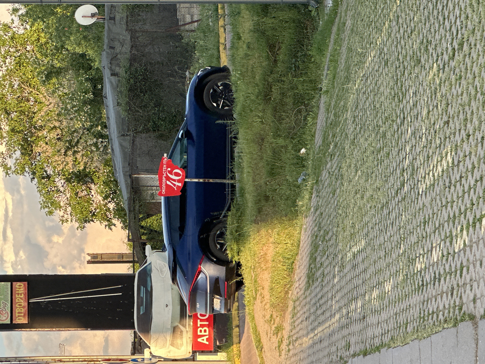 The image captures a street scene in Sofia, Bulgaria, dominated by parked cars on a paved surface interspersed with grass. A white vehicle is positioned alongside a dark blue car, with a red sign reading "АВТО" (car) visible. Adjacent to the blue car is a sign, possibly advertising a commercial establishment, featuring the number "46." A sign above the white vehicle reads "ОТВОРЕНО" (open). Behind the parked cars, a grassy area and some greenery indicate a roadside verge. In the background, a building or structure is visible against the sky. The general atmosphere is urban with elements of greenery.