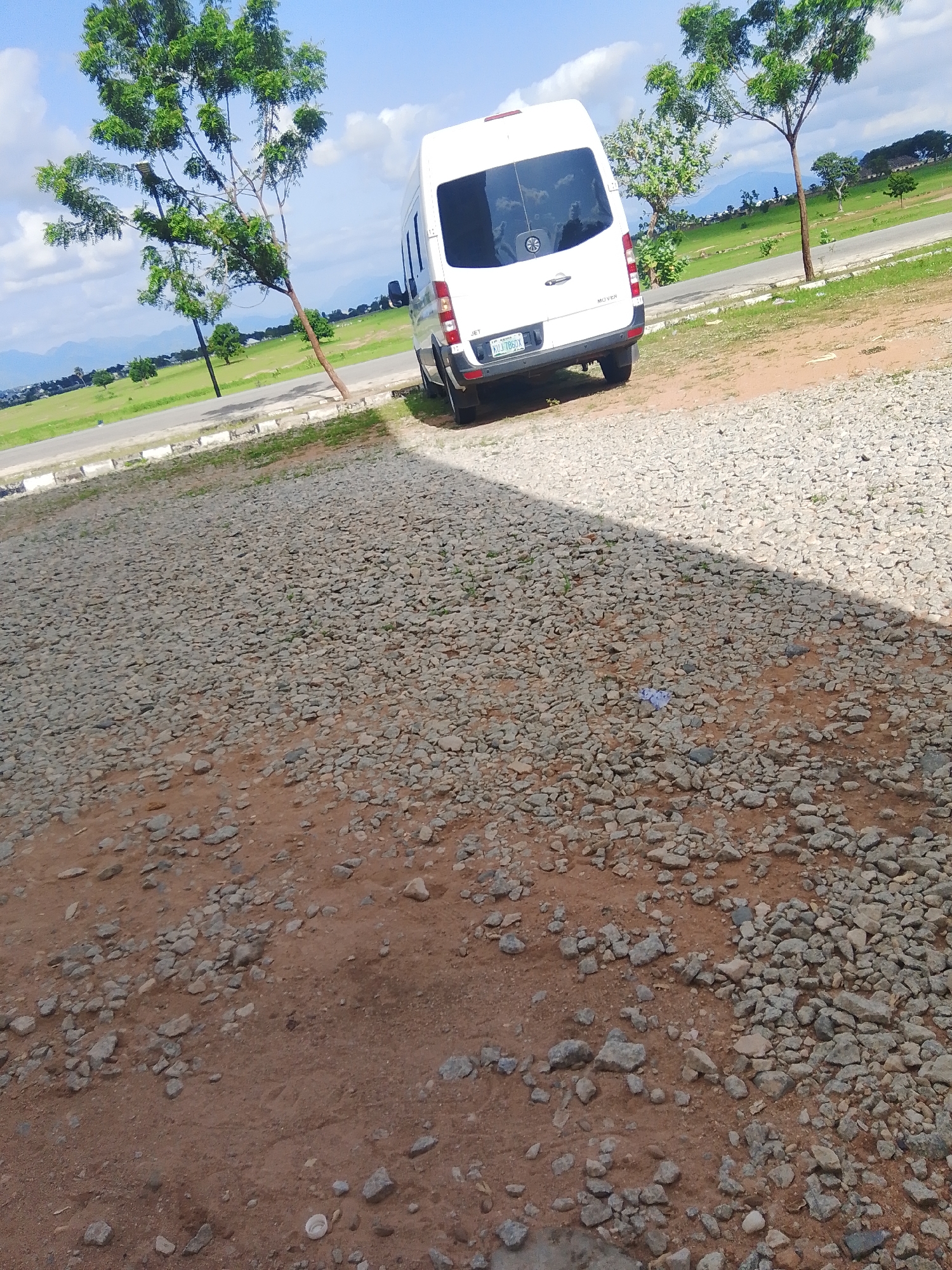 A white Mercedes-Benz Sprinter van is parked on a gravel lot, facing away from the camera. The van's license plate is visible and reads "KUJ 786DX". The scene appears to be outdoors during the day, with a bright, partly cloudy sky. In the background, there are trees and a grassy field, with mountains visible in the distance. The area is rocky and dusty, with some sparse vegetation growing amongst the stones. A shadow from a tree or structure falls across the right side of the image. The overall setting suggests a semi-arid or rural environment, possibly in Jalingo, Nigeria, given the location context. There are no people visible in the image.