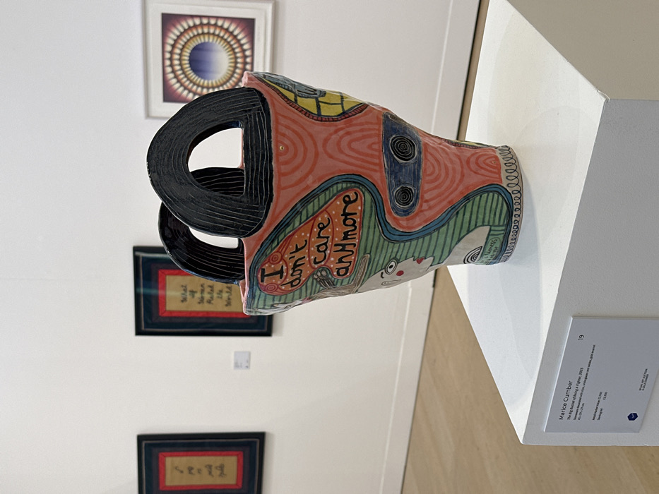 This image depicts an interior view of an art gallery or exhibition space, identifiable as Sotheby's in London, United Kingdom, likely hosting an exhibition by The Koestler Arts Trust. The central subject is a multi-colored ceramic sculpture, "The Big Bucket of Being A Fighter" by Marice Cumber, created in 2023. The irregularly shaped vessel, resembling a bucket or vase with two black, arch-shaped handles, is adorned with various patterns, including concentric swirls, stripes, and dots, in shades of pink, green, blue, yellow, and black. A stylized face with red lips is visible on one side, and the phrase "I don't care anymore" is prominently inscribed in orange lettering on a red background. The sculpture, made of stoneware with slips, underglazes, oxides, and gold enamel, rests on a white rectangular plinth in the foreground. An accompanying white plaque details the artwork's information, including its dimensions (40 x 31 x 27 cm) and retail market value (£2,100). The background consists of plain white walls and a light-colored wooden floor. Three other framed artworks are visible on the far wall: two to the left feature text on a reddish-brown background (one reading "What if Women Ruled the World"), and one to the right displays a concentric circular abstract design in vibrant blues, purples, yellows, and reds. No individuals or active interactions are present in the scene.
