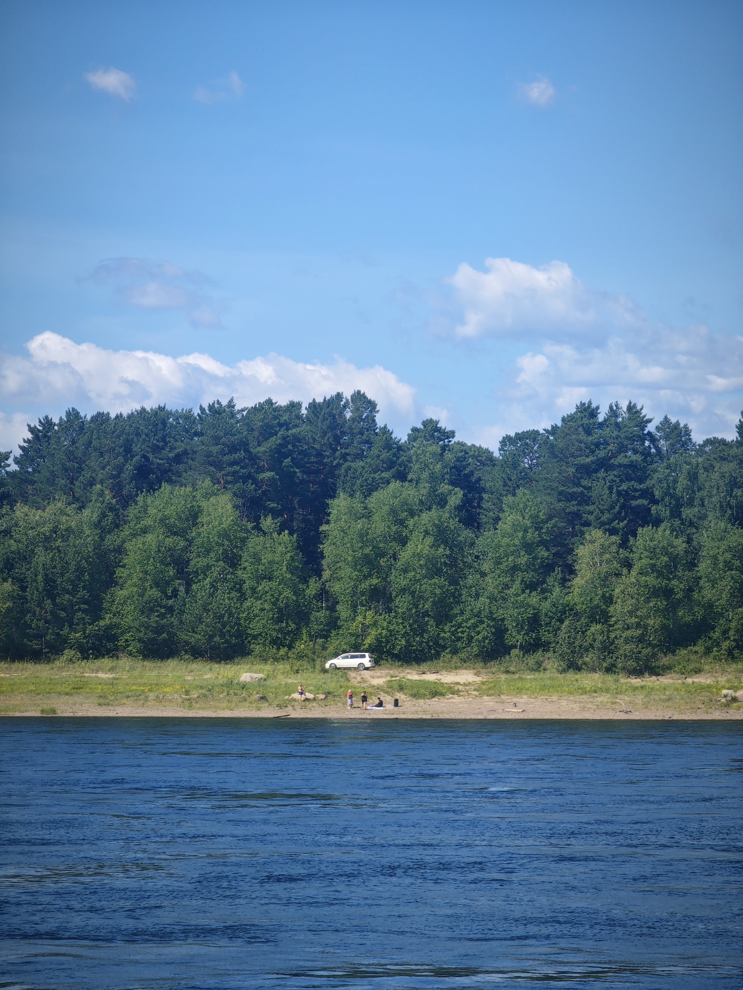 The image depicts a serene natural landscape with a wide, calm blue river in the foreground. The river stretches across the lower half of the frame, its surface textured by gentle ripples. In the middle ground, a sandy shore separates the river from a dense, lush green forest that dominates the upper portion of the image. The forest is a mix of deciduous and coniferous trees, with a varied canopy creating a rich tapestry of greens. A few scattered rocks are visible on the shore.
On the sandy shore, near the edge of the forest, a white van is parked. A small group of people, appearing to be enjoying the outdoors, are scattered along the shoreline. One person is standing near the water's edge, while others are seated or lying down, seemingly relaxing on the sand. The sky above is a bright, clear blue with a few scattered white cumulus clouds, suggesting a pleasant, sunny day. The overall impression is one of a peaceful, natural setting perfect for recreation, likely a riverside park or a natural area accessible by road, such as one might find near the settlement of Абалаково, Russia, given its proximity to the Yenisei River.