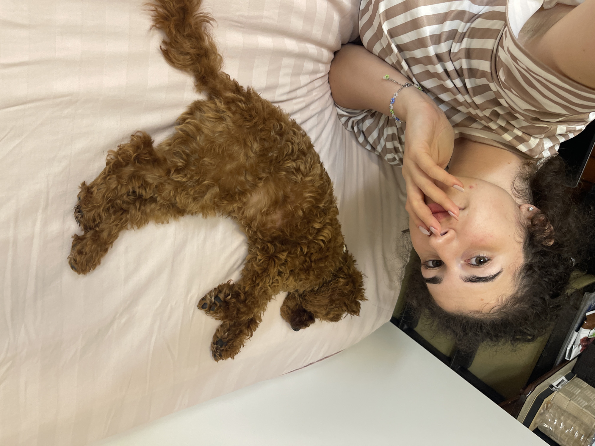 The image depicts an overhead selfie taken in an indoor setting, identified by the prompt as Sofia, Bulgaria. A young female individual and a small dog are sharing a bed.
The female is situated in the lower right portion of the frame, lying on her back with her head oriented towards the lower left. She has dark, curly hair, dark eyes, and a light complexion. She is wearing a brown and white striped top. Her left hand is raised, with fingers gently resting near her mouth and chin. A silver chain bracelet adorned with small blue and green beads is visible on her right wrist, and a small stud earring is in her visible earlobe. Her fingernails are painted with a light-colored polish. She is looking directly towards the camera.
Above and to the left of the female, in the upper-middle section of the frame, a small, reddish-brown, curly-haired dog, consistent with a Poodle or Poodle mix, is lying on its back on the bed. Its belly is exposed, and its paws are curled, indicating a relaxed or sleeping state. The dog's head is towards the lower left, and its tail extends towards the upper right.
The bed is covered with light pink or beige sheets that feature subtle, horizontal stripe patterns. The sheets appear lightly wrinkled. To the far left and at the bottom edge of the frame, a white vertical surface (possibly a wall or headboard) and a darker, indistinct area containing miscellaneous items (e.g., packaging, textiles) are visible, characteristic of a bedroom environment. The scene is illuminated by soft, even lighting.