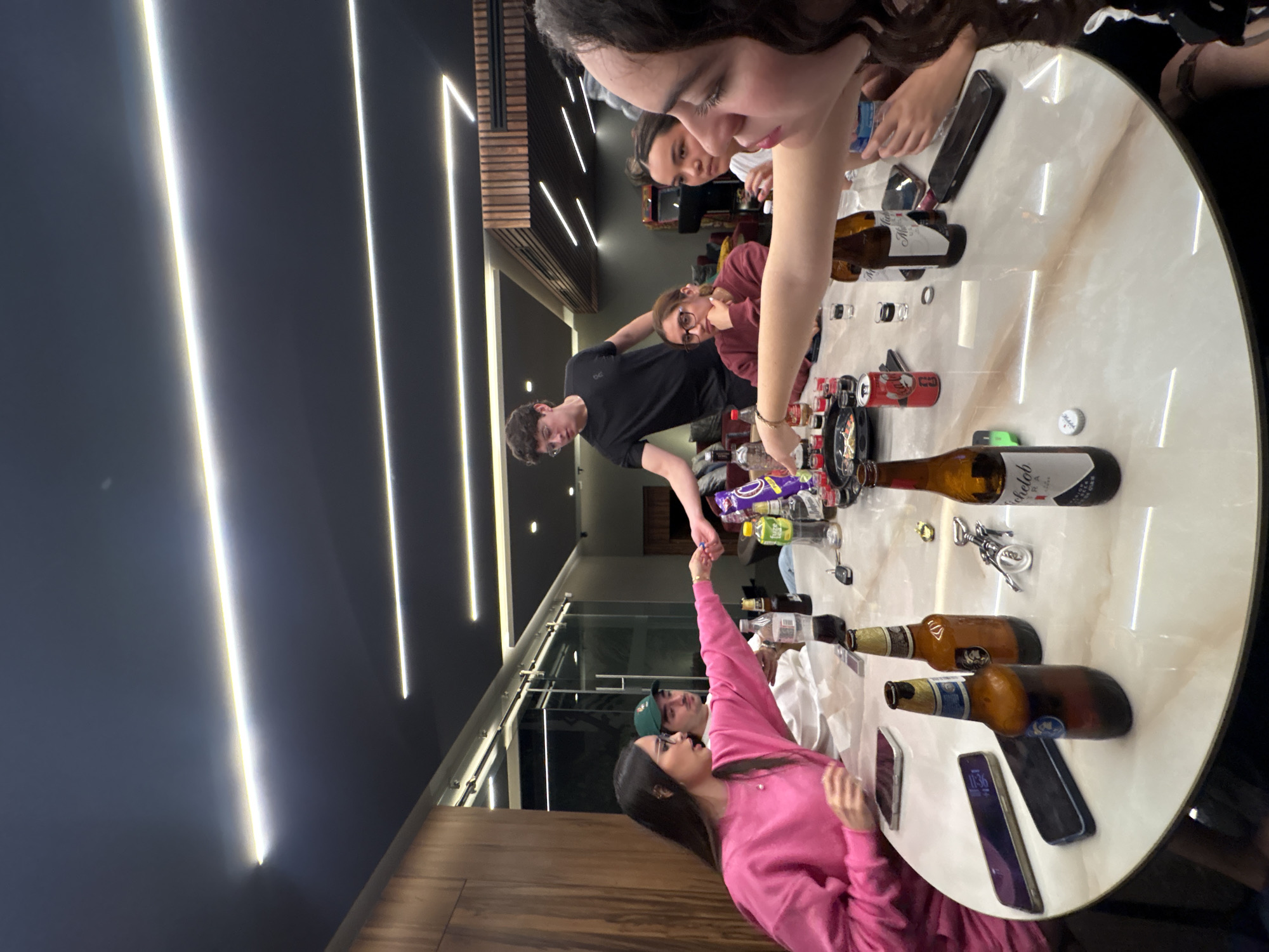 The image depicts an indoor social gathering of at least seven young adults around a large, round, light-colored table, likely in San Bernardino Tlaxcalancingo, Mexico. **People:** - On the left, a young woman with long dark hair, wearing a bright pink sweatshirt and glasses, extends her right arm across the table, holding a small, dark object towards the center. Her left hand rests on the table near a smartphone. - Behind her, a young man with a mustache and a green baseball cap wears a white t-shirt and looks towards the table. - Standing in the left-center background, a young man with short dark hair and glasses, wearing a black t-shirt, also reaches towards the table with his left arm. - On the right side of the table, a young woman with dark hair and glasses, in a maroon shirt, rests her chin on her hand, observing the activity. - Next to her, a partially obscured young woman with dark hair pulled back, wearing a white shirt, also looks towards the table. - In the immediate foreground on the right, a young woman with long, curly dark hair, wearing a dark top, has her right arm extended across the table, participating in the central interaction. Her face is in profile. - Other individuals are partially visible in the background. **Objects and Interactions:** - The large, round table has a reflective, light-colored surface with subtle marbling. - Numerous items are scattered across the table, including: multiple empty or partially full beer bottles (Modelo Especial, Michelob Ultra), several small shot glasses (some with clear liquid, others empty), a red Coca-Cola can, a bottle opener/corkscrew, a loose bottle cap, at least four smartphones, a green soda bottle (Sprite/7 Up brand), a clear plastic water bottle, a purple bag of "Takis Fuego" brand snacks, small black dishes containing various items, and a set of keys. - The central interaction involves the woman in pink and the woman in the foreground extending their arms, suggesting an exchange or presentation of an item at the center of the table. **Environment:** - The room features a dark grey/black ceiling with multiple parallel, linear LED light fixtures emitting a bright, warm white light, which is prominently reflected on the table surface. - The walls consist of light brown wooden paneling on the left and dark walls in the background. - A large glass partition or sliding door system