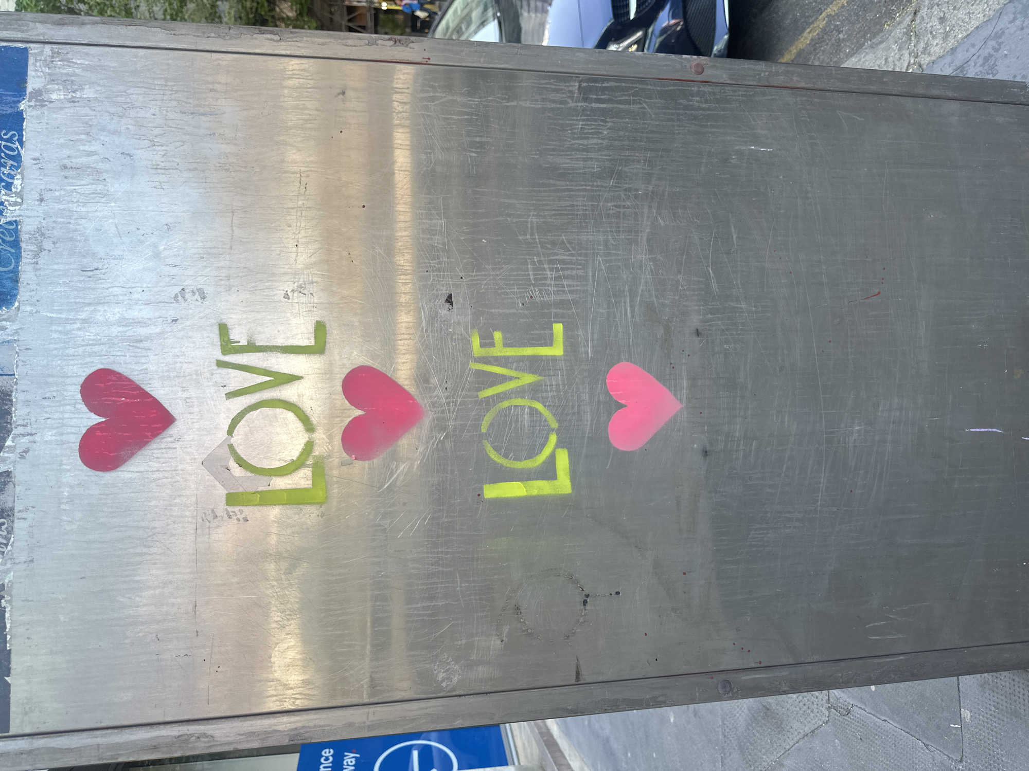 A close-up, medium-angle shot reveals a scratched, metallic surface, possibly a public utility box, upon which the word "LOVE" is rendered twice. Each instance features a bright green, stencil-like font for the letters "L," "O," and "V." In place of the letter "E" are vivid pink heart shapes. The placement suggests an urban location, potentially London, United Kingdom. The image is angled vertically, with the ground and partial view of a vehicle visible at the top right corner. The style suggests street art or graffiti.