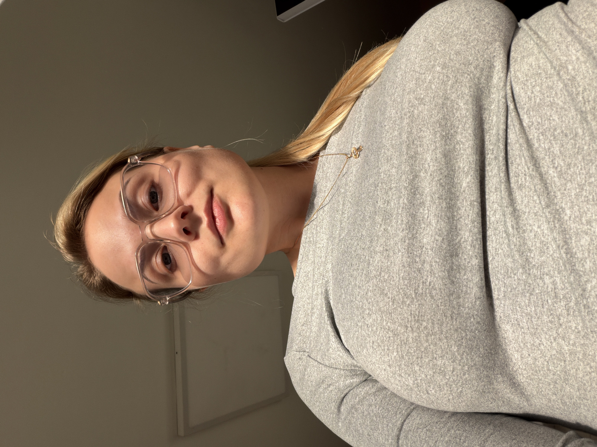 This low-angle portrait features a young woman with blonde hair, looking upwards directly into the camera with a gentle, slight smile. She wears modern, clear-framed glasses that sit prominently on her face. Her blonde hair, styled back, is visible over her right shoulder (as oriented in the image). She is dressed in a light grey, long-sleeved top or sweatshirt, which appears to be made of a soft, textured fabric. A delicate gold-colored necklace with a small, indistinct pendant is visible around her neck.
The background is simple and uncluttered, primarily a plain light grey wall on the left side of the frame, with a darker, undefined area suggesting a corner or another surface on the right. A thin white vertical line, possibly a door frame or architectural trim, is faintly visible in the lower left background. The lighting is soft and warm, casting a gentle glow on her face, particularly on her forehead and cheek, and highlighting strands of her hair. This illumination suggests an indoor setting, possibly with natural light from a window or a bright overhead source. Her expression is calm and friendly. No text is discernible in the image. While the provided context indicates Tel-Aviv, Israel, no specific visual cues within the image confirm this location.